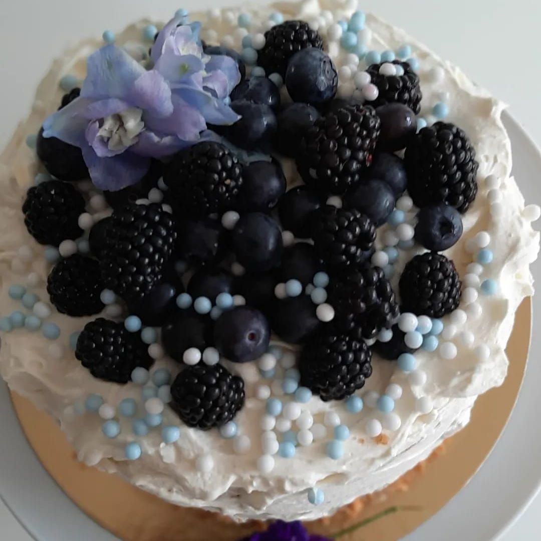 Taart met witte glazuur, versierd met bramen, bosbessen, blauwe hagelslag en een blauwe bloem.