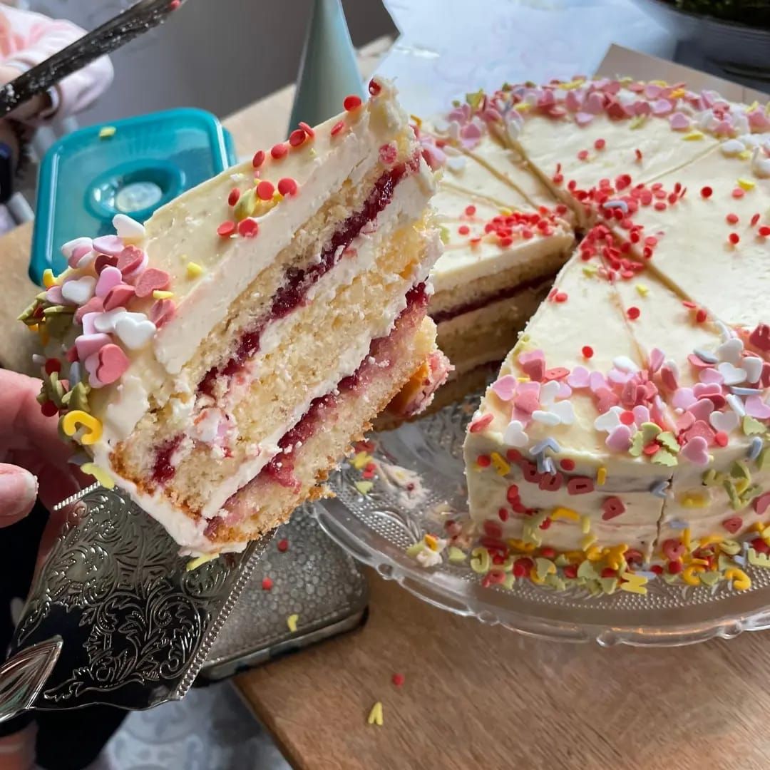 Een stuk geglazuurde cake met roze hagelslag en jamvulling, dat van een transparant bord wordt getild.