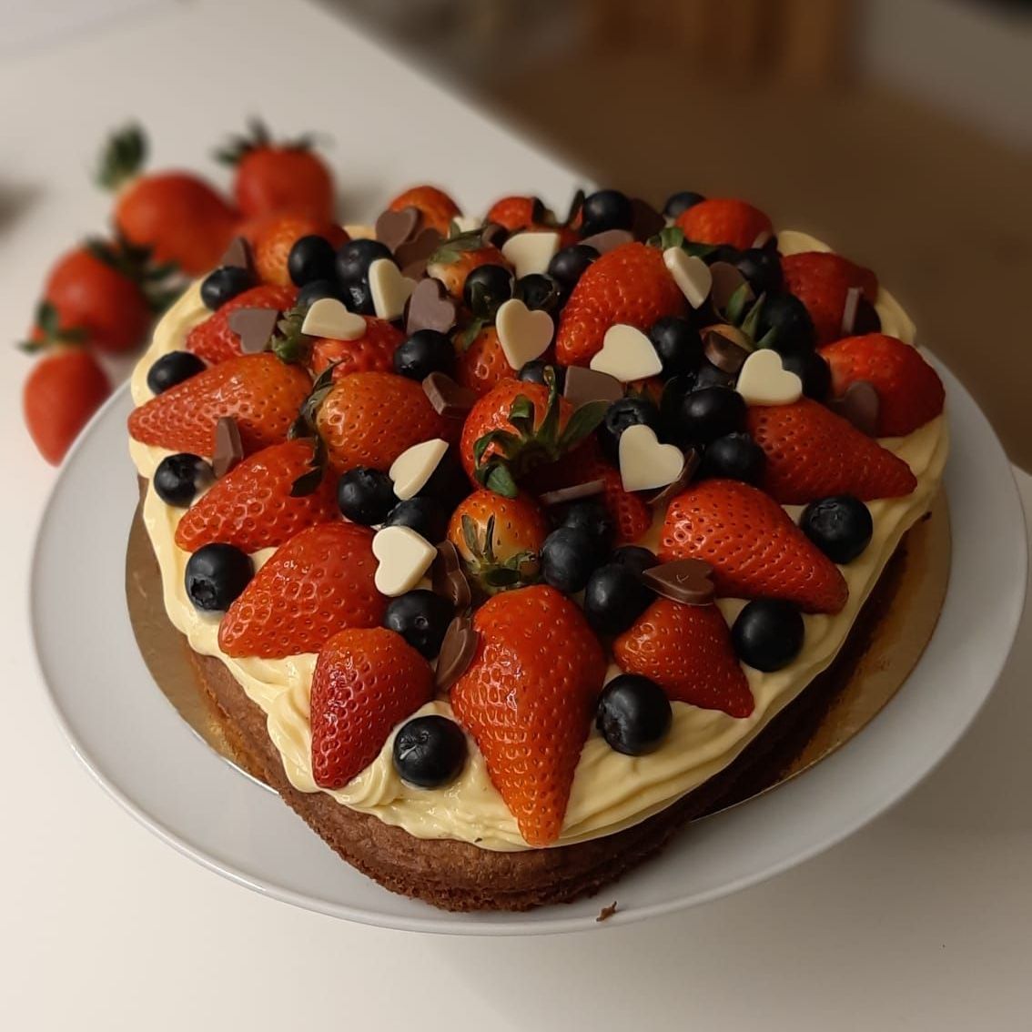Hartvormige taart met aardbeien, bosbessen en hartjes van witte chocolade op een bord, met aardbeien op de achtergrond.