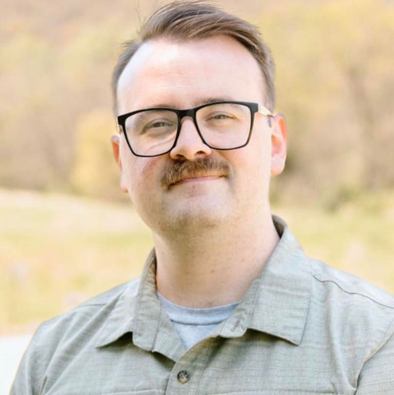 A person with a mustache and glasses wearing a collared shirt, smiling gently in front of a blurred outdoor field.