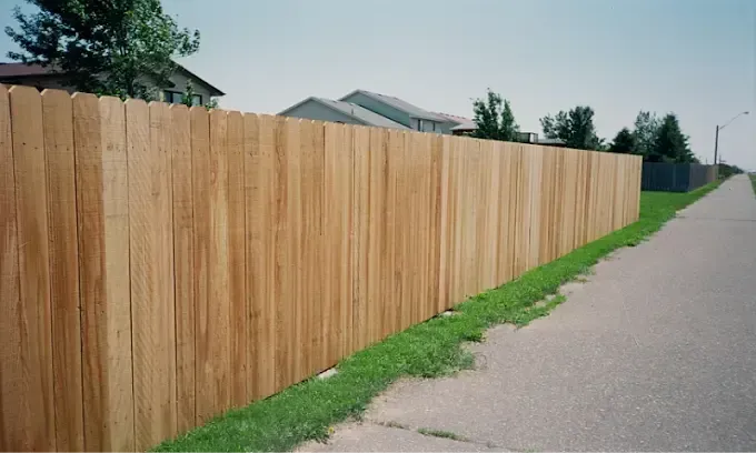 A tall, new wooden fence runs along the edge of a sidewalk next to a grassy area, with houses visible in the distance.