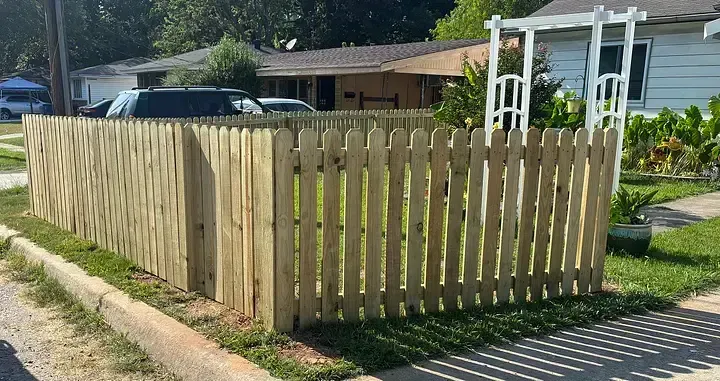 A newly installed wooden picket fence surrounds a front yard, featuring an arched white trellis near a suburban house.