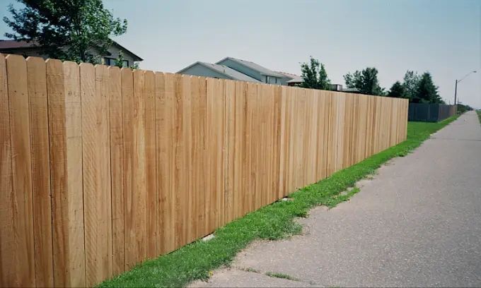 A tall, light-brown wood privacy fence runs alongside a paved sidewalk next to a grassy patch.