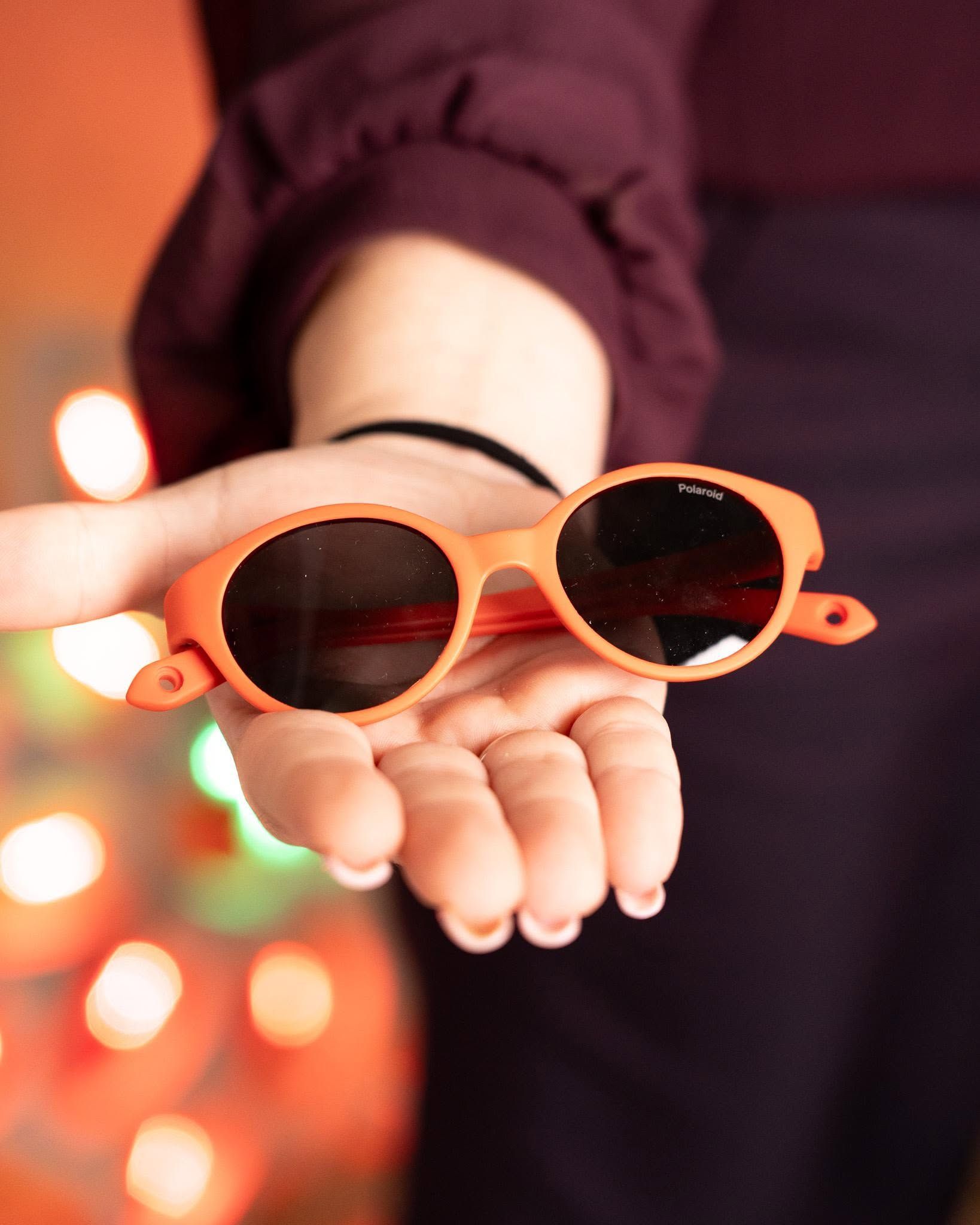 Hand holding orange sunglasses with blurred warm lights in the background