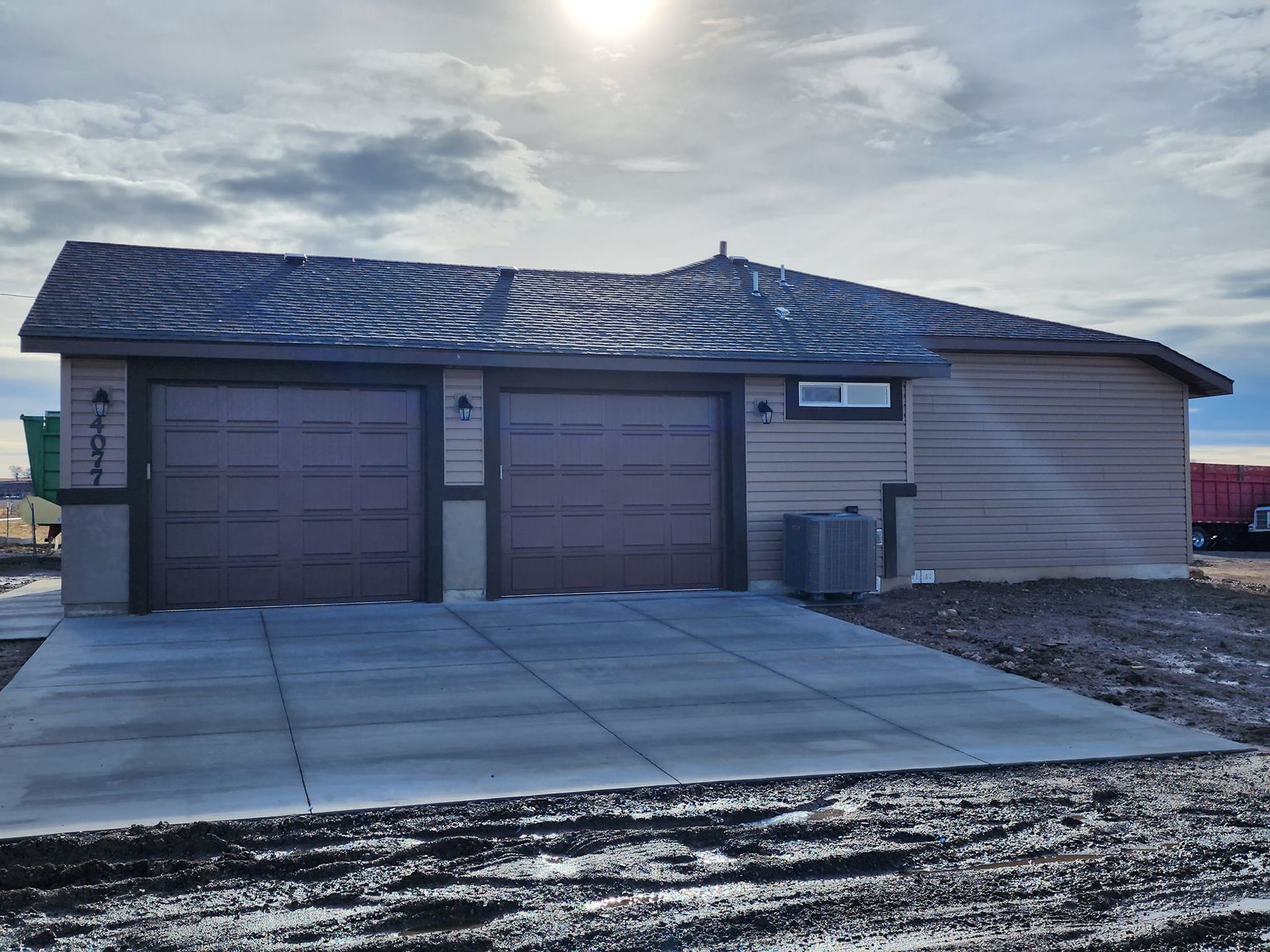 Tan-sided house with two-car garage, brown doors, concrete driveway, cloudy sky, and the sun.