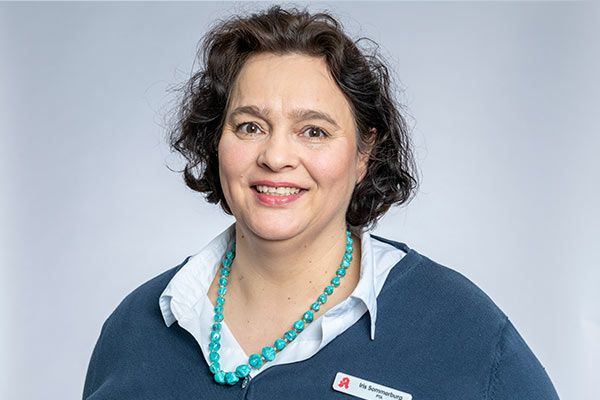 Eine Frau in einem blauen Pullover und einer türkisfarbenen Halskette lächelt in die Kamera.