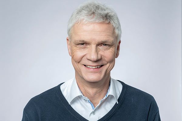 Ein Mann in einem blauen Pullover und einem weißen Hemd lächelt in die Kamera.