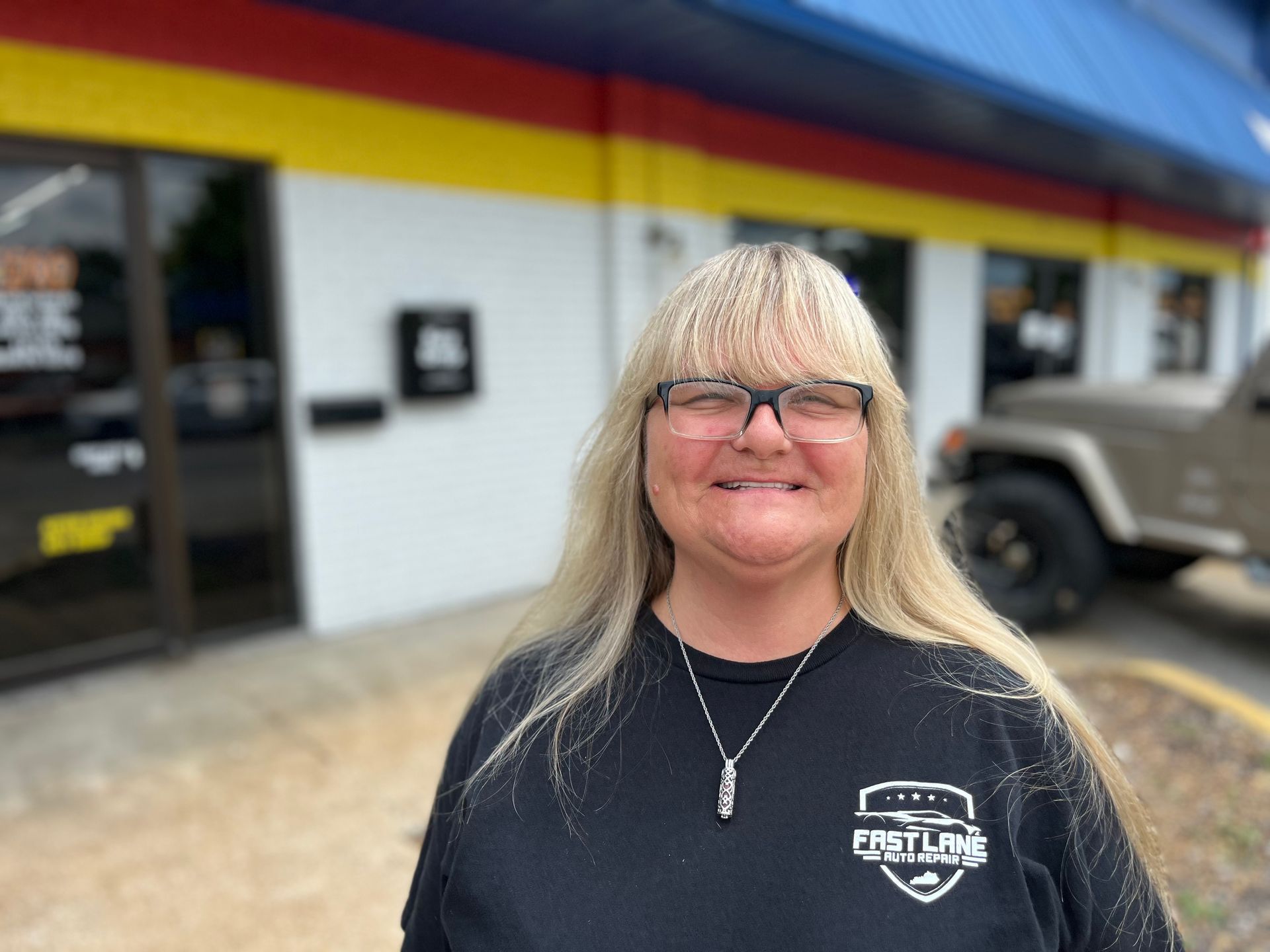 Woman with blonde hair and glasses smiles in front of a building with red, yellow, and blue stripes.
