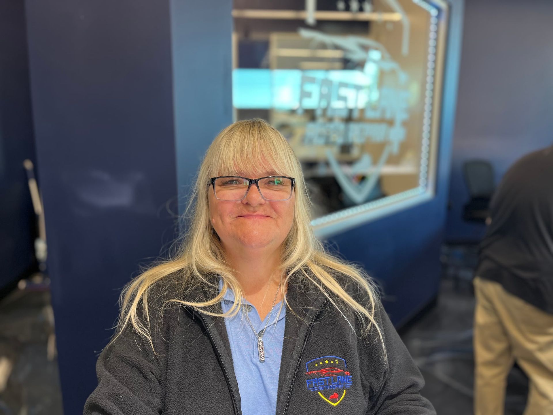 Woman with blonde hair and glasses smiles in front of a building with red, yellow, and blue stripes.