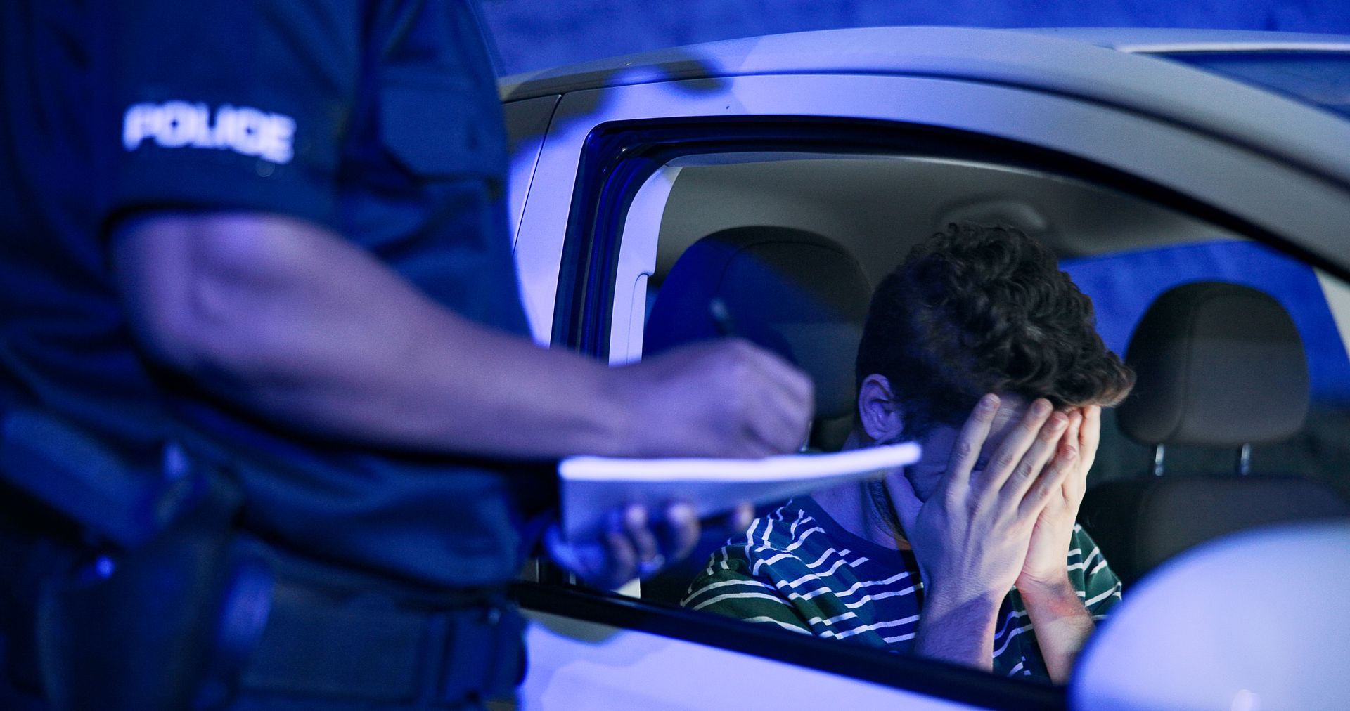 Police officer writing a ticket for a driver. Driver inside car covers face, appearing distressed. Night setting.