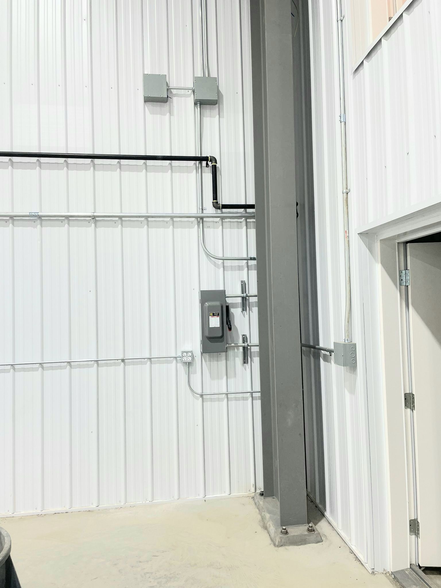 Interior view of a white corrugated metal wall with gray metal support beam, electrical conduits, and a disconnect switch.