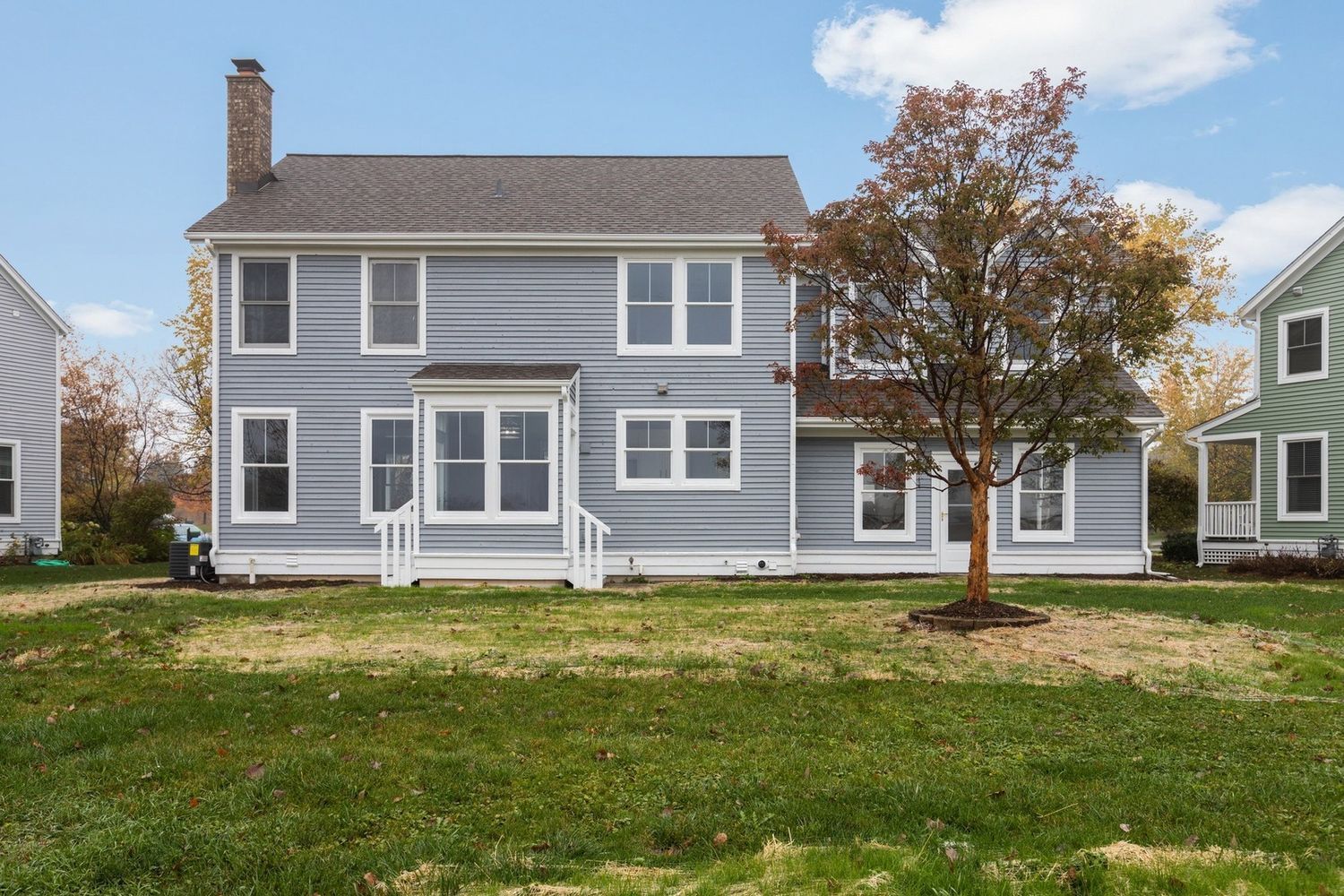 Two-story blue house with white trim, chimney, and a tree on a green lawn under a blue sky.