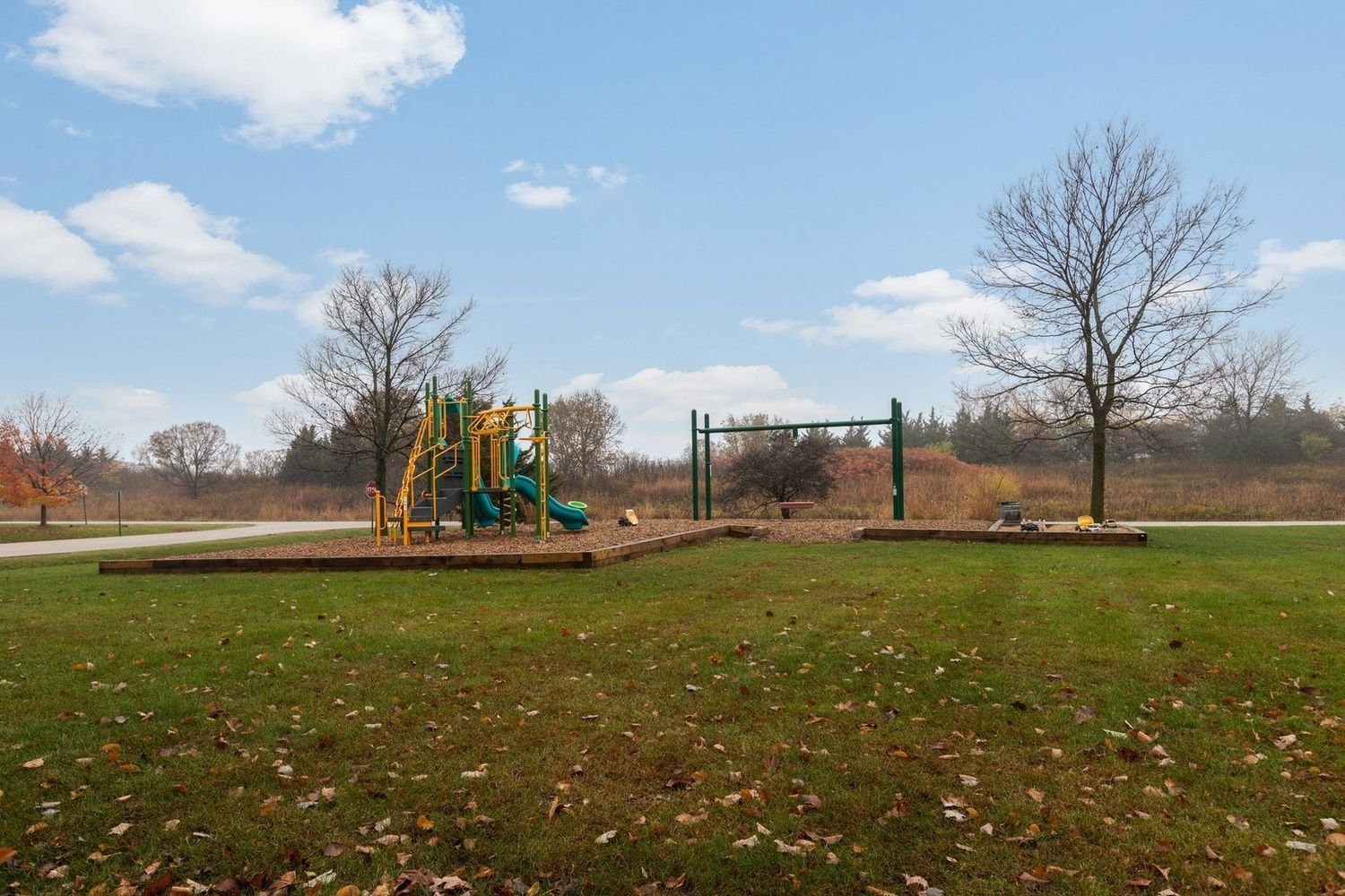 Playground in a park with swings and a slide, set against a blue sky with scattered clouds.
