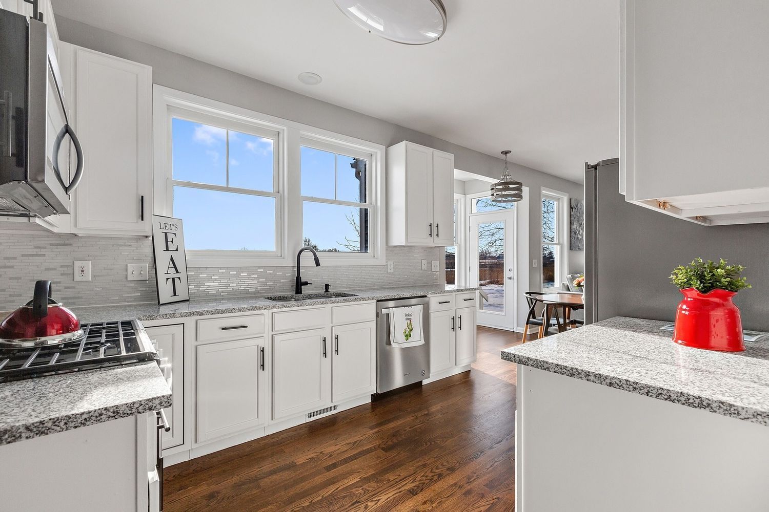 White kitchen with dark wood floors, white cabinets, and granite countertops; sunlight streams through windows.