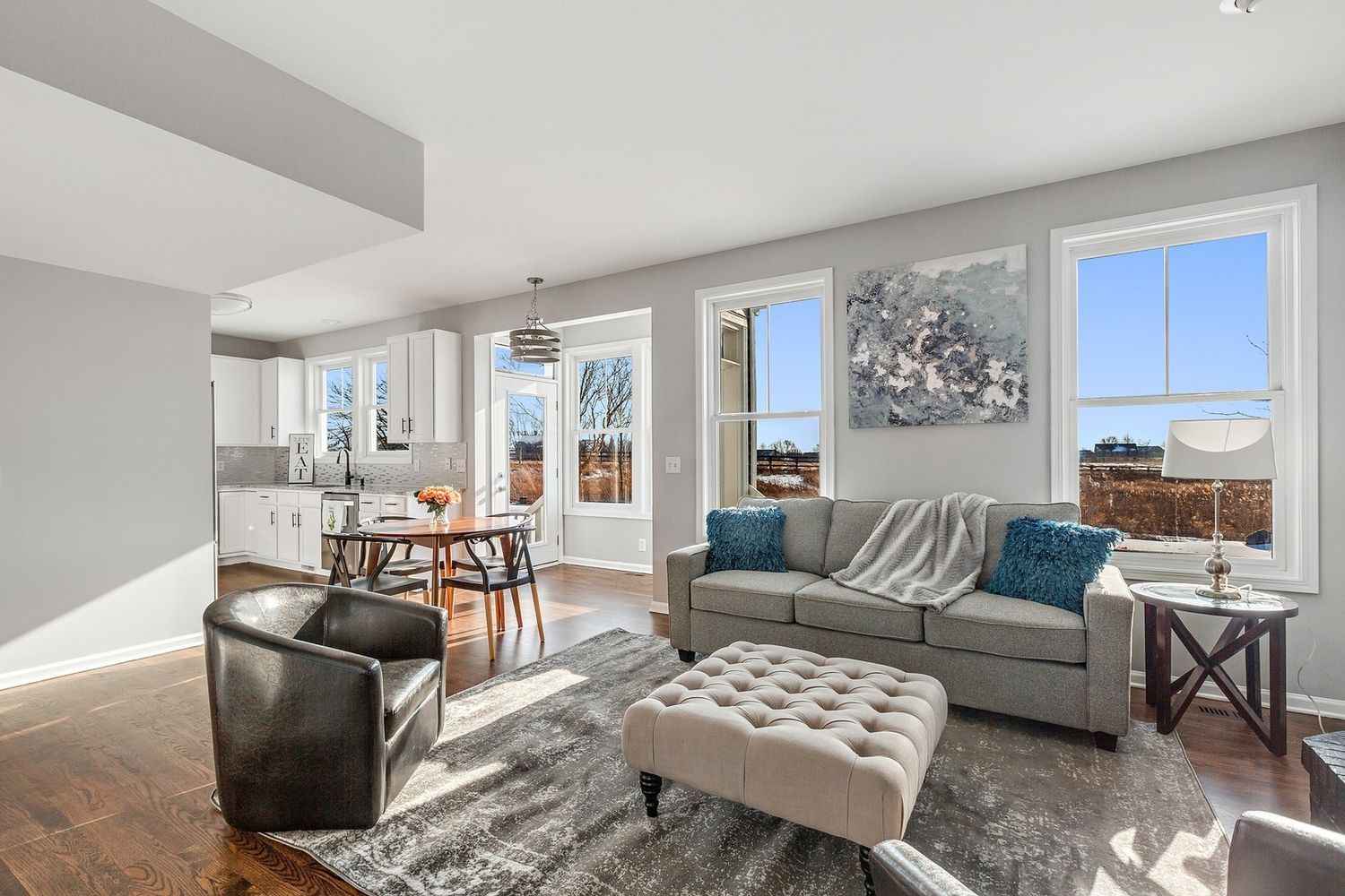 Open living room with gray sofa, ottoman, and dark wood floors; view of kitchen and windows.