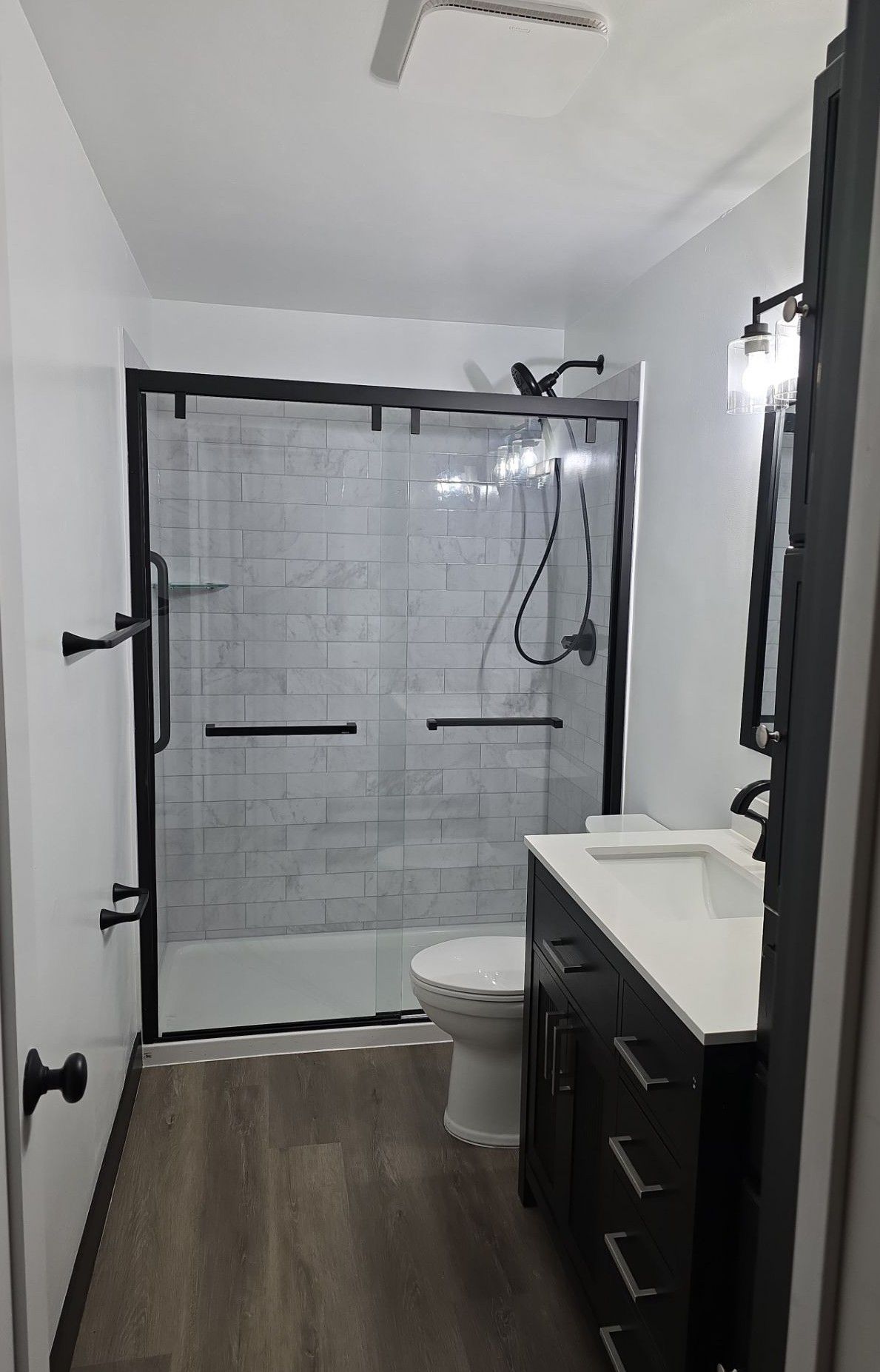 A modern bathroom featuring a black-framed glass walk-in shower, a dark vanity, white walls, and wood-look flooring.