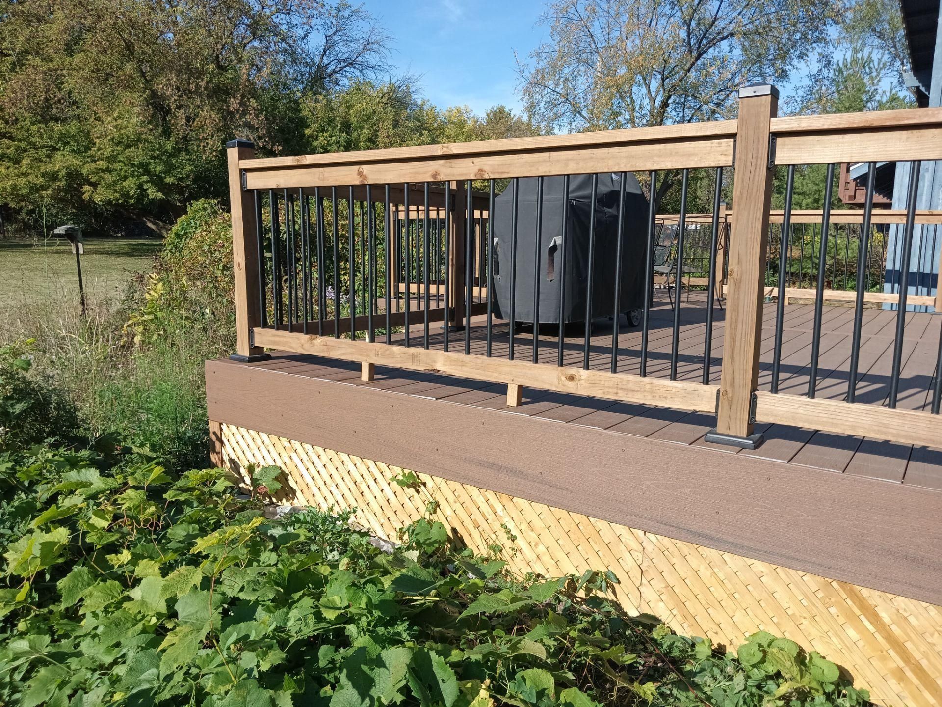 A wooden deck with a black metal railing and lattice skirting, surrounded by green foliage on a sunny day.