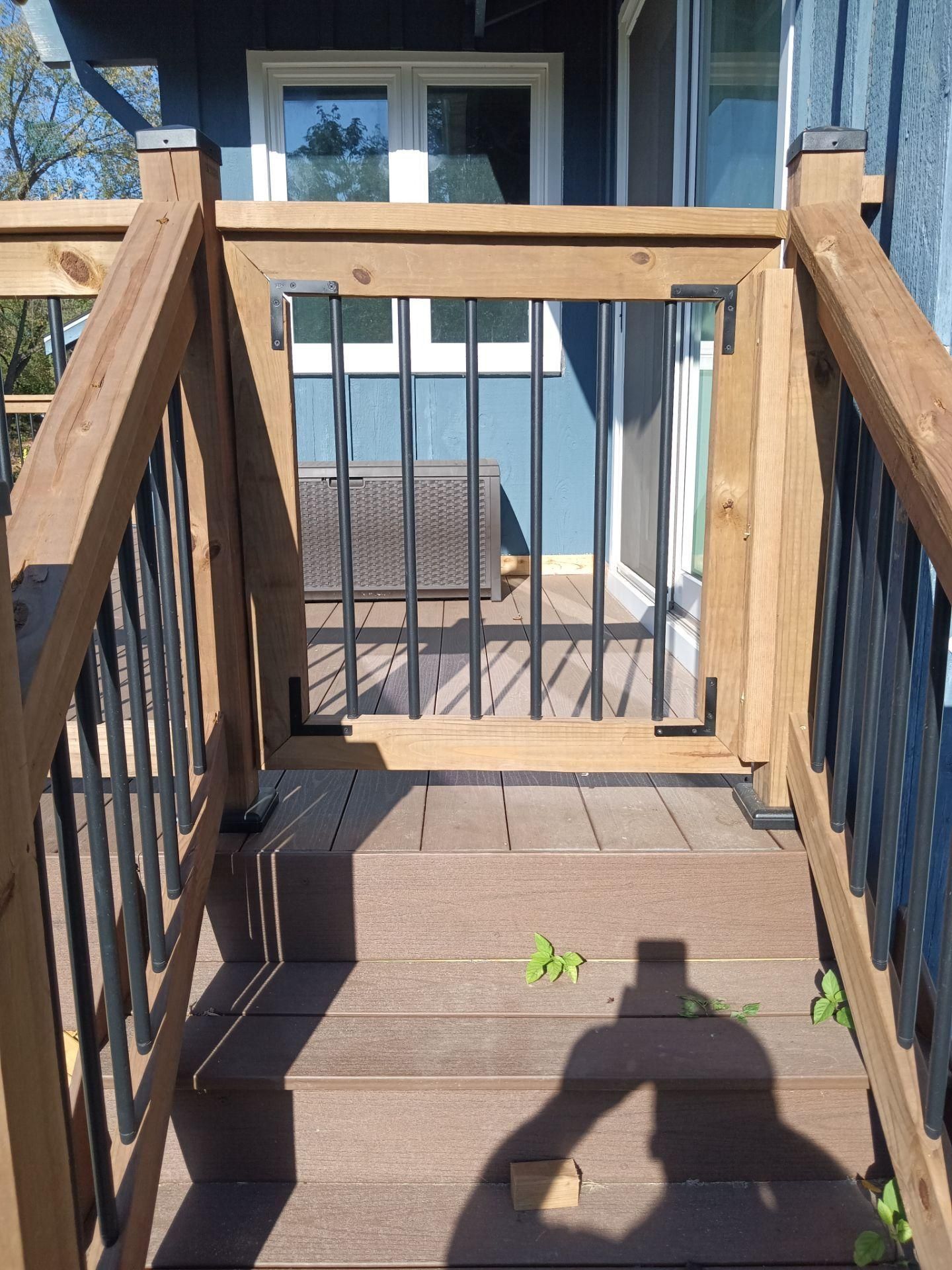 A wooden deck gate with black metal balusters installed at the top of a set of composite stairs on a house exterior.