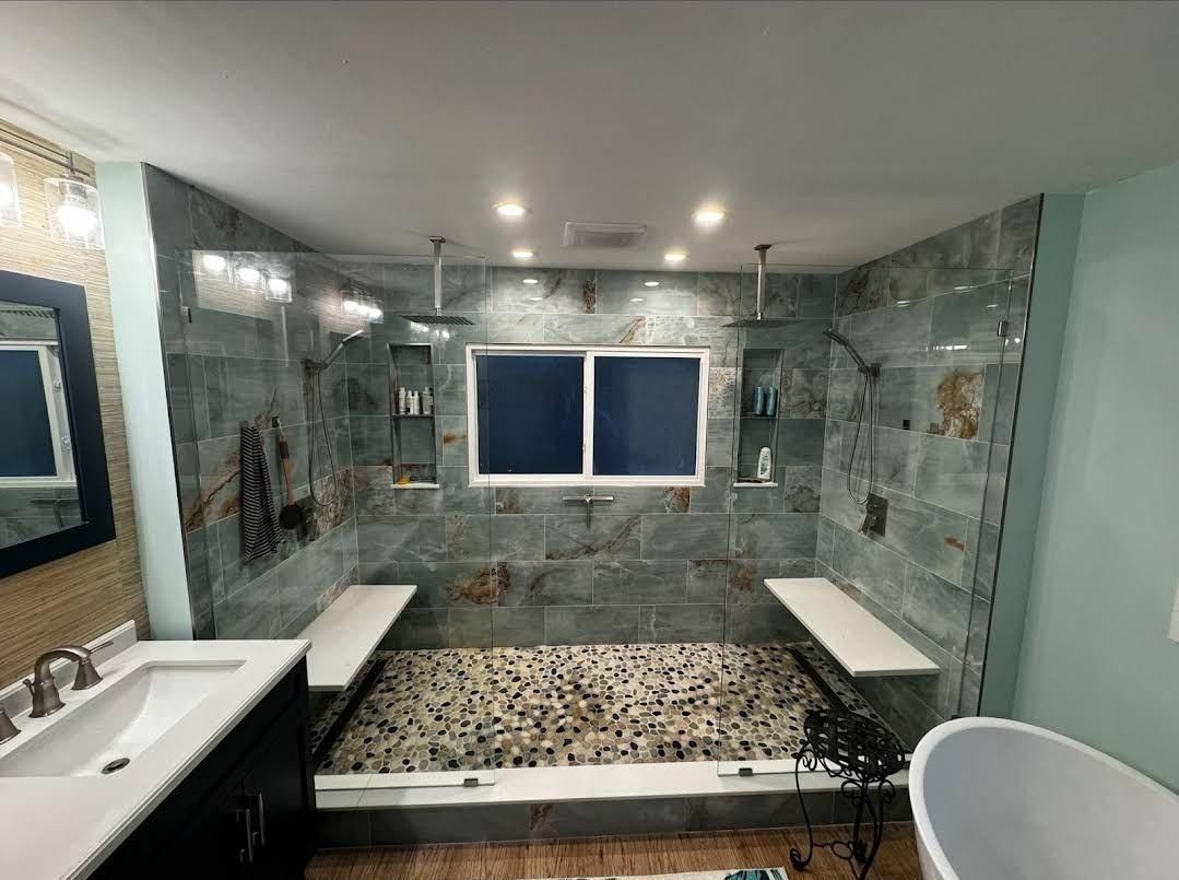 A walk-in shower with stone tile walls, a pebble floor, two white benches, and a central window with recessed niches.