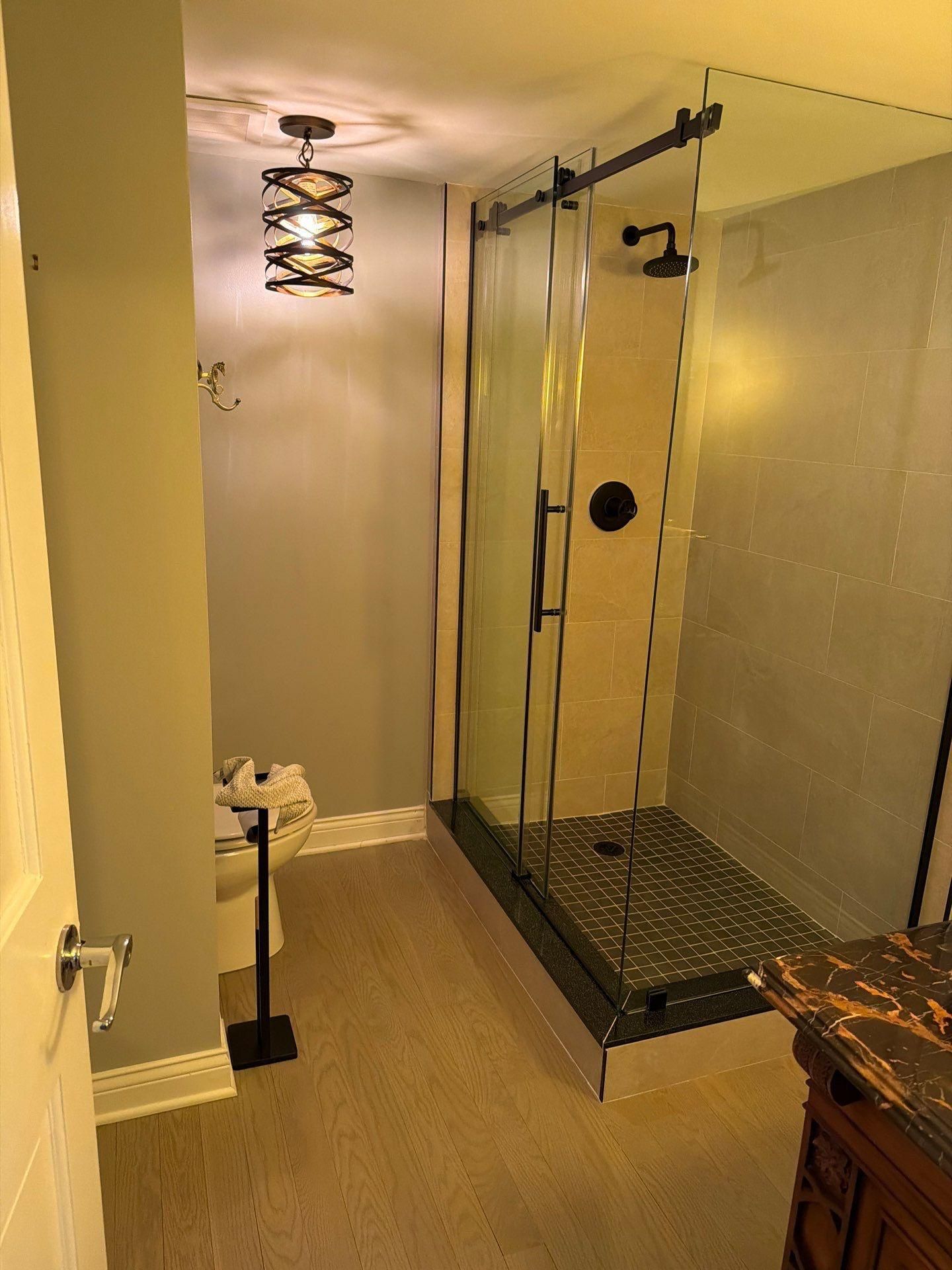 A bathroom with a glass-enclosed shower, a toilet with a stand, grey walls, wood-look flooring, and a hanging light fixture.