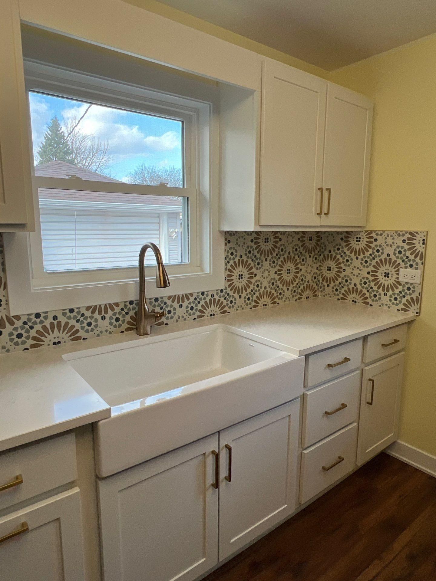 A farmhouse kitchen sink with a bronze faucet and patterned backsplash below a window with white cabinets and wood floors.