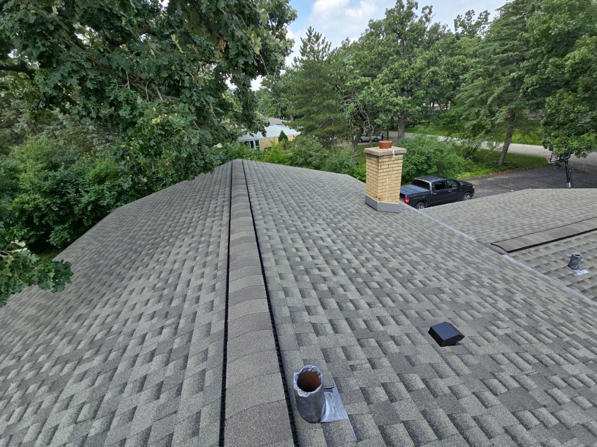 An elevated view of a gray asphalt shingle roof featuring a central ridge vent, a brick chimney, and a small black vent.