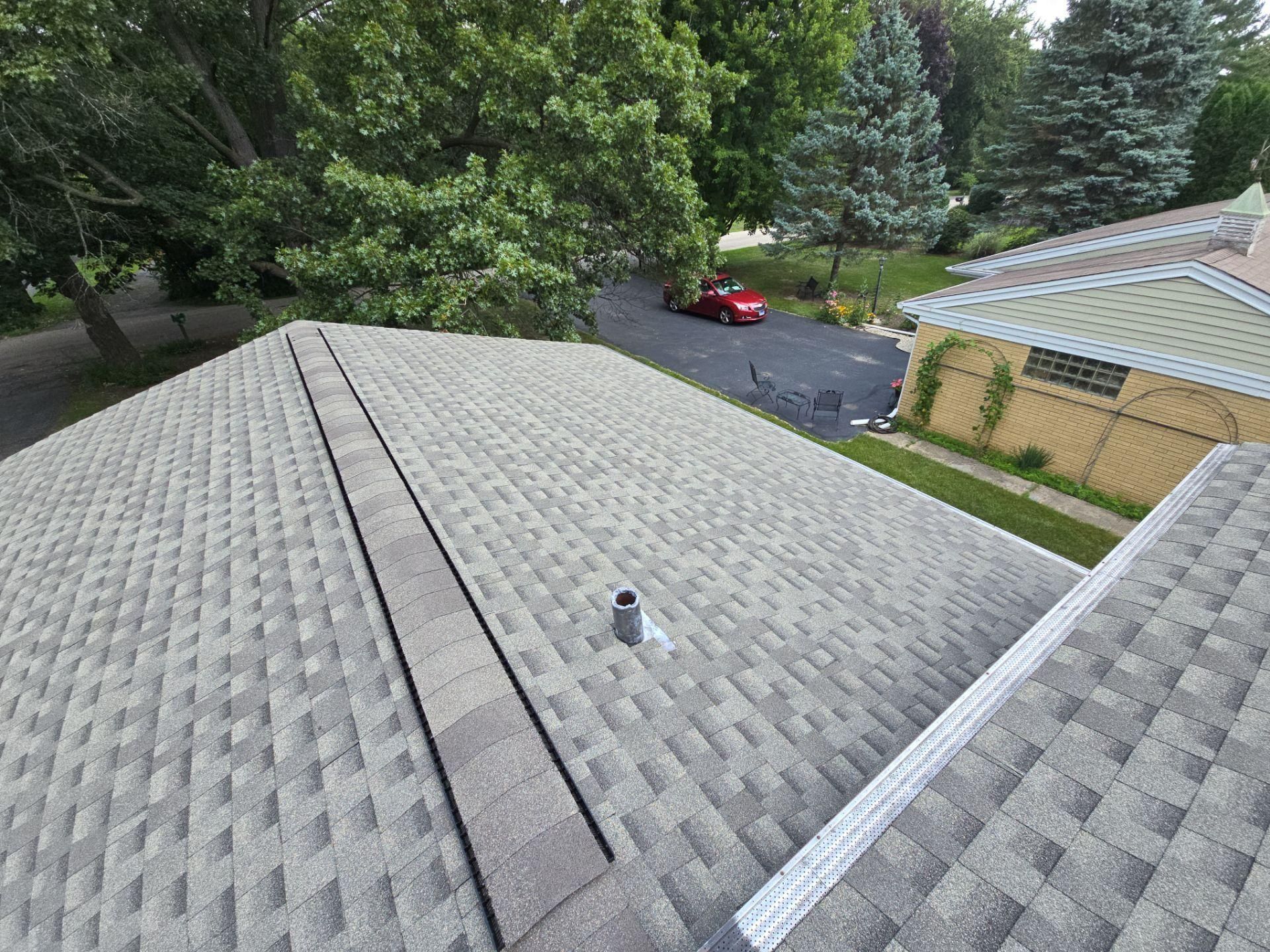 An elevated, slightly angled view of a textured gray shingled roof with a single vent pipe, overlooking a yard and road.