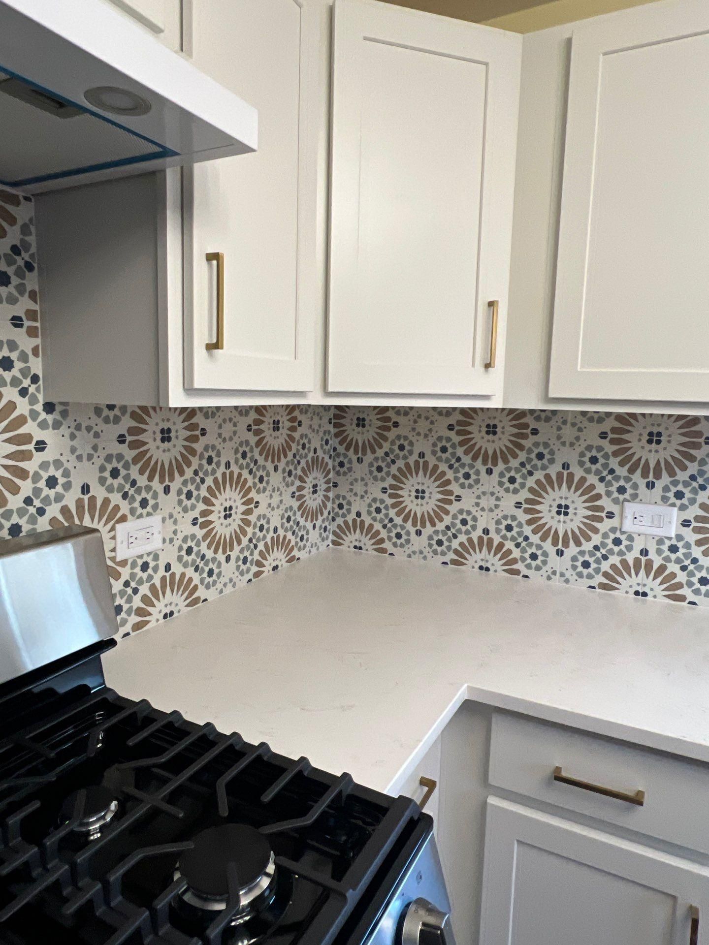 A corner of a kitchen with a gas stove, white cabinets, gold handles, and a patterned tile backsplash.