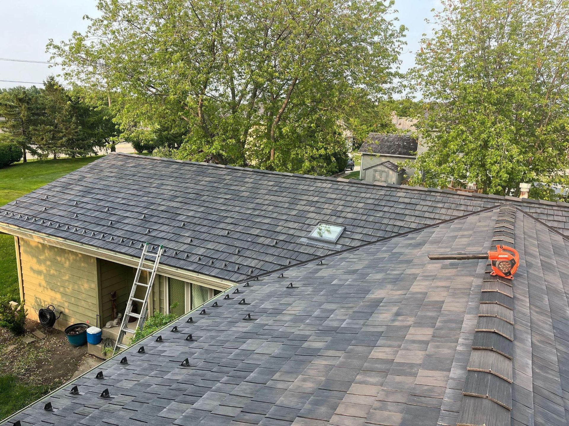 A high-angle view of a grey shingled roof under construction with an orange blower, a ladder, and trees in the background.