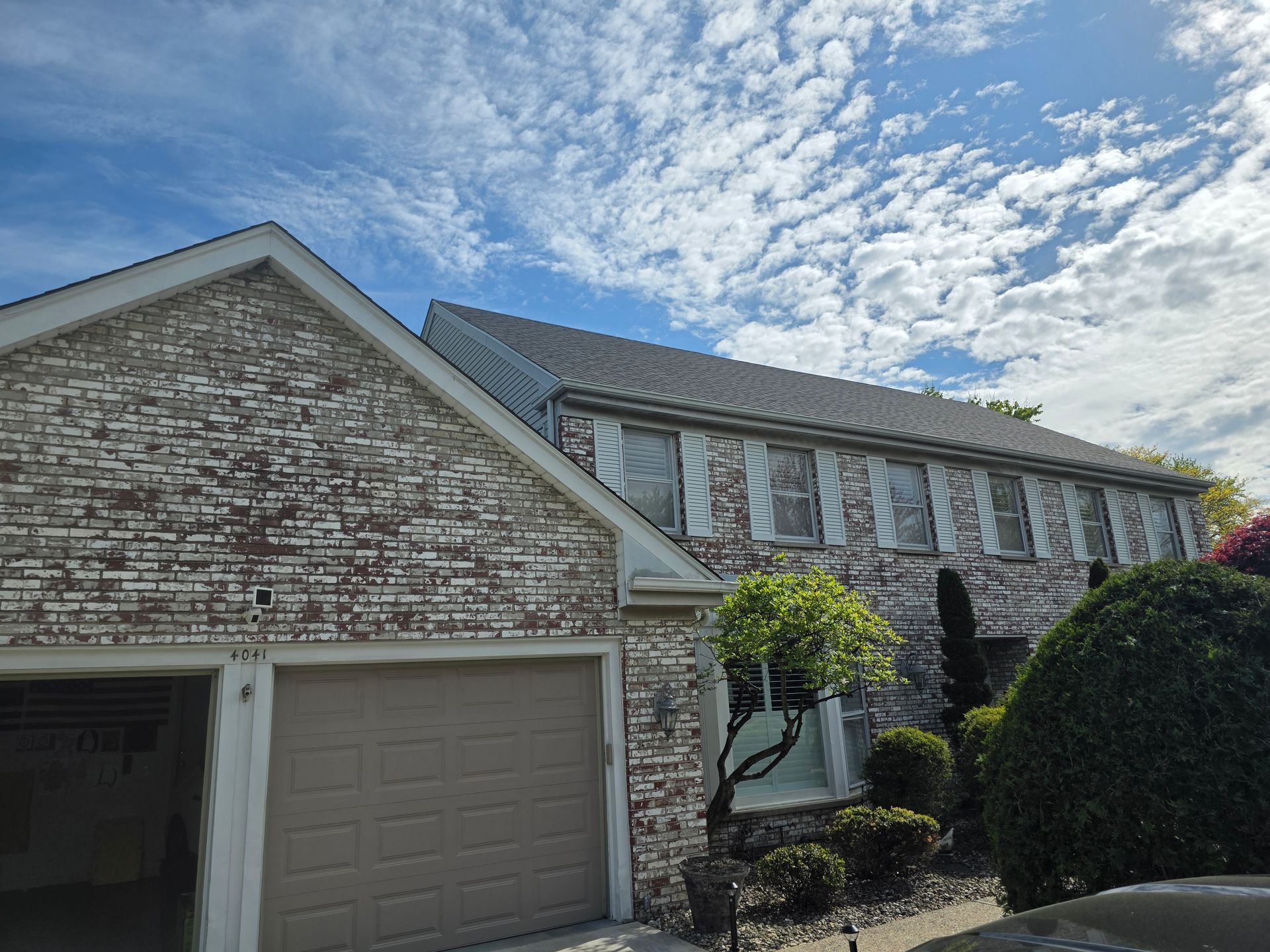 A tan brick, two-story house with a garage, dark roof, and white shutters under a blue sky with scattered clouds.