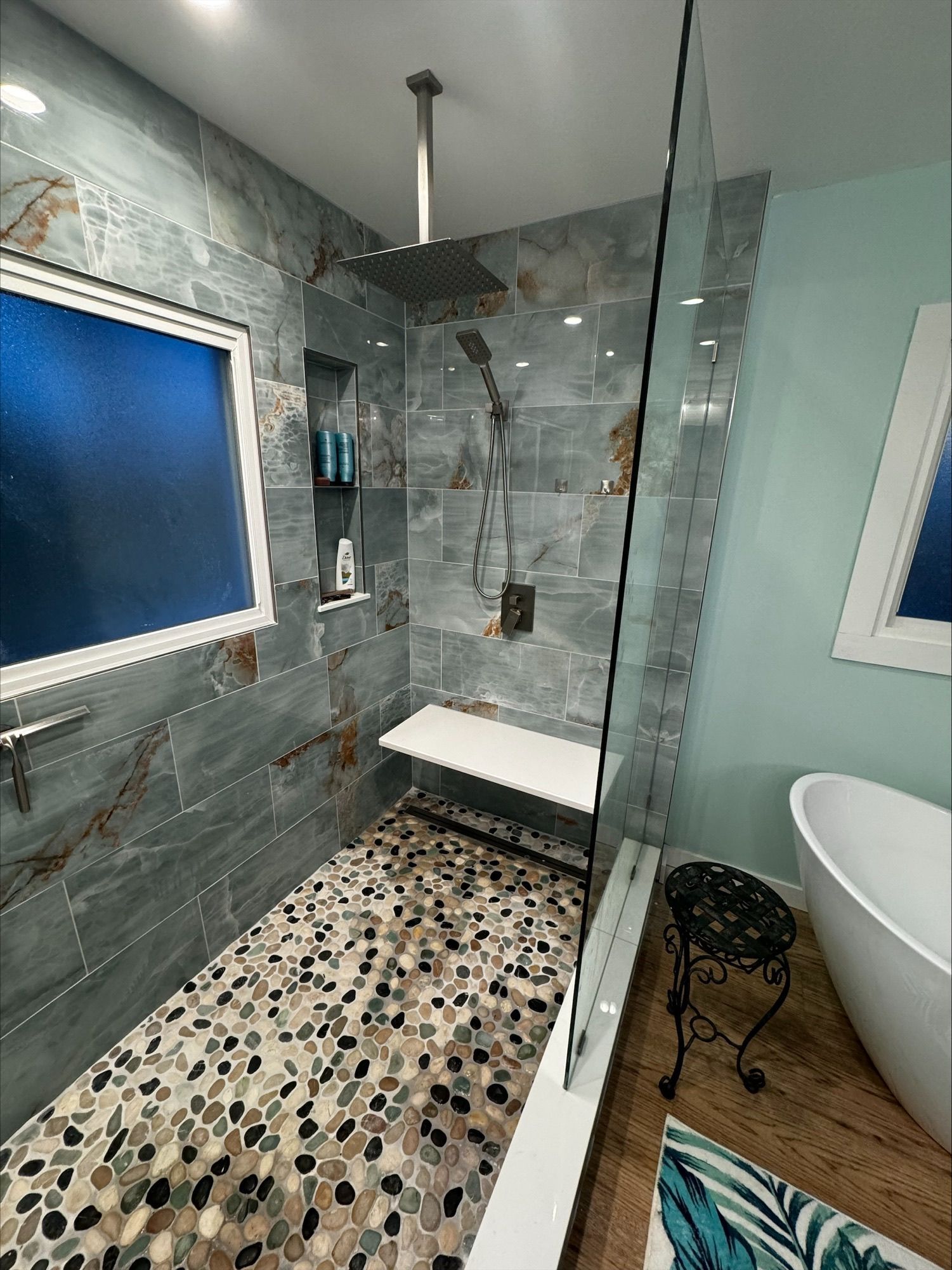 A modern shower with blue-grey stone tiles, a pebble floor, a built-in bench, and a glass partition next to a bathtub.