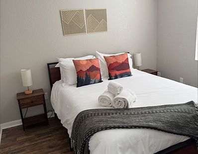 Una cama hecha con dos cojines con estampado de paisajes al atardecer, sábanas blancas y una manta gris oscuro entre dos mesitas de noche.