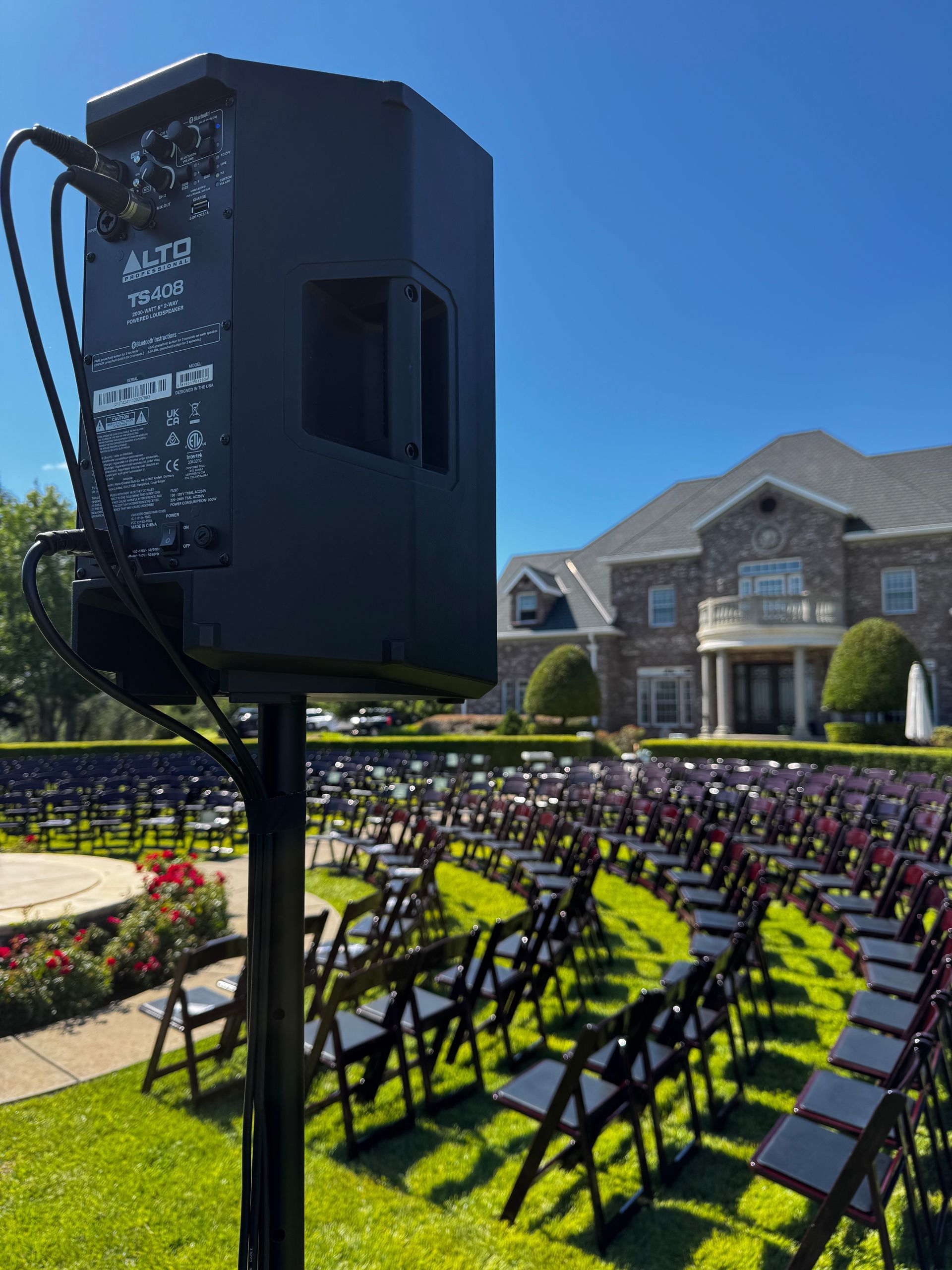 Outdoor wedding ceremony setup with rows of black chairs on green lawn and a large speaker in front of a stone building