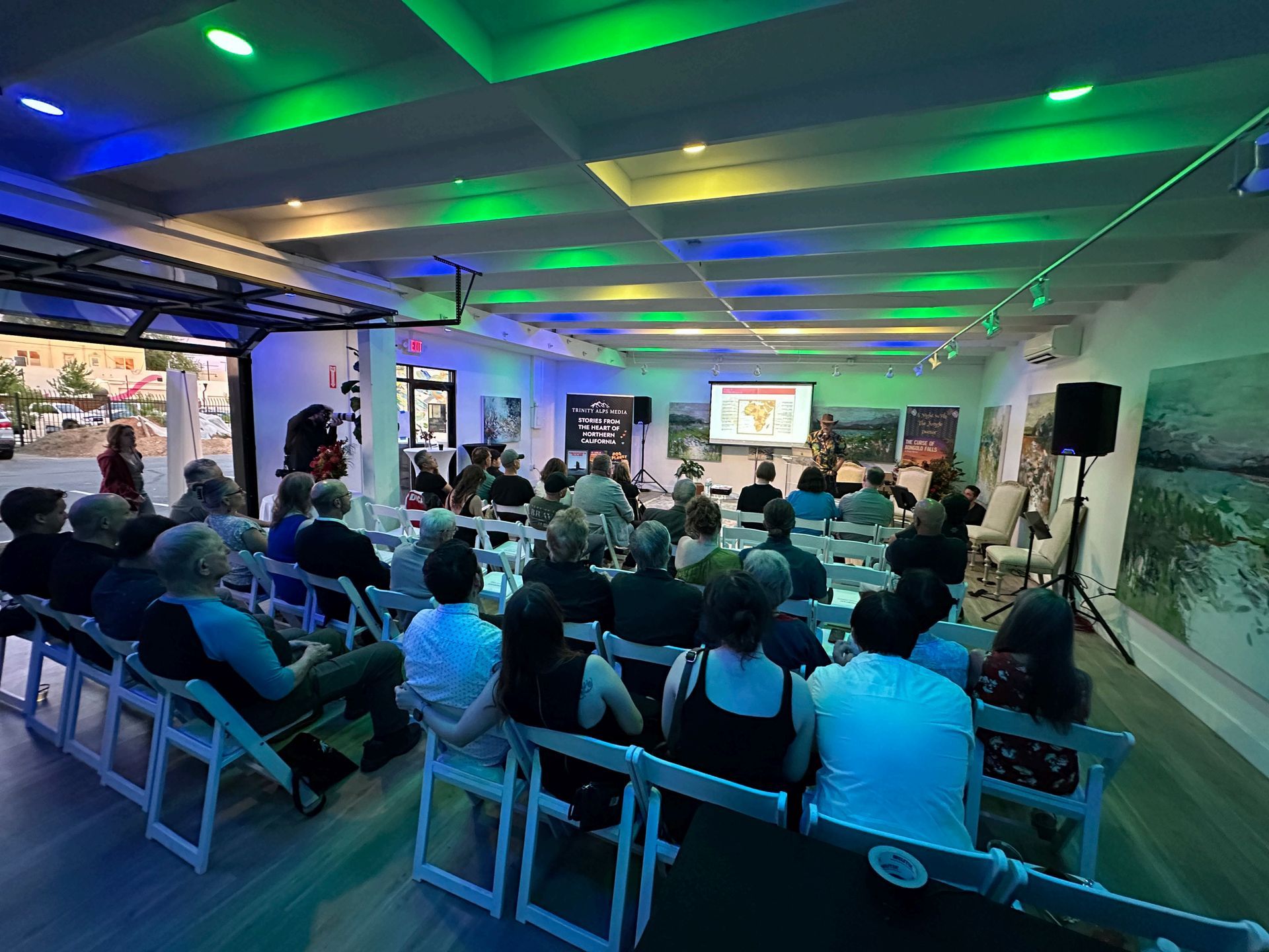 Audience seated in a modern conference room, watching a presentation on a large screen under green-blue lighting