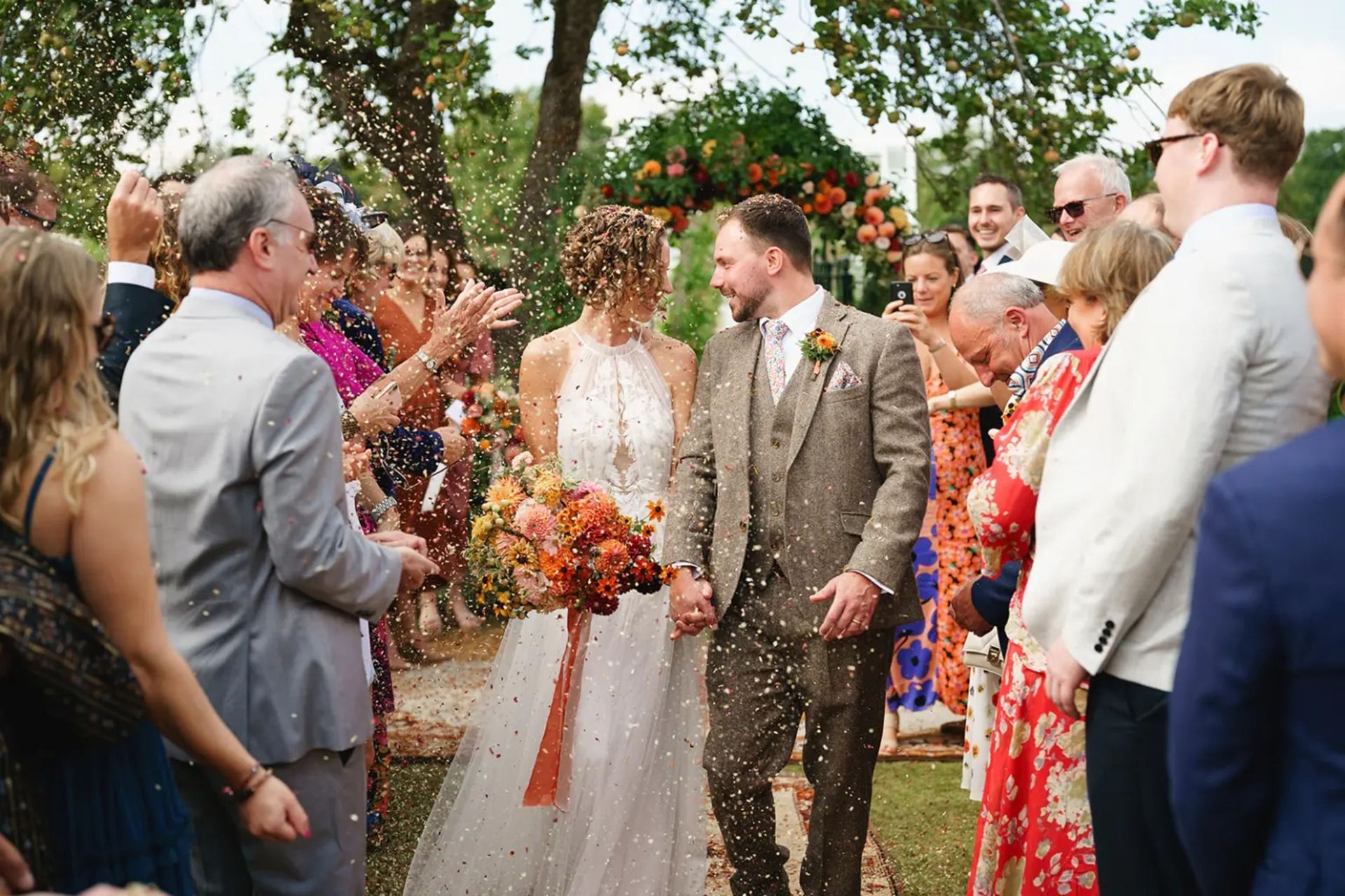 Wedding couple standing in an outdoor aisle as guests applaud, with colorful floral decor and trees in the background