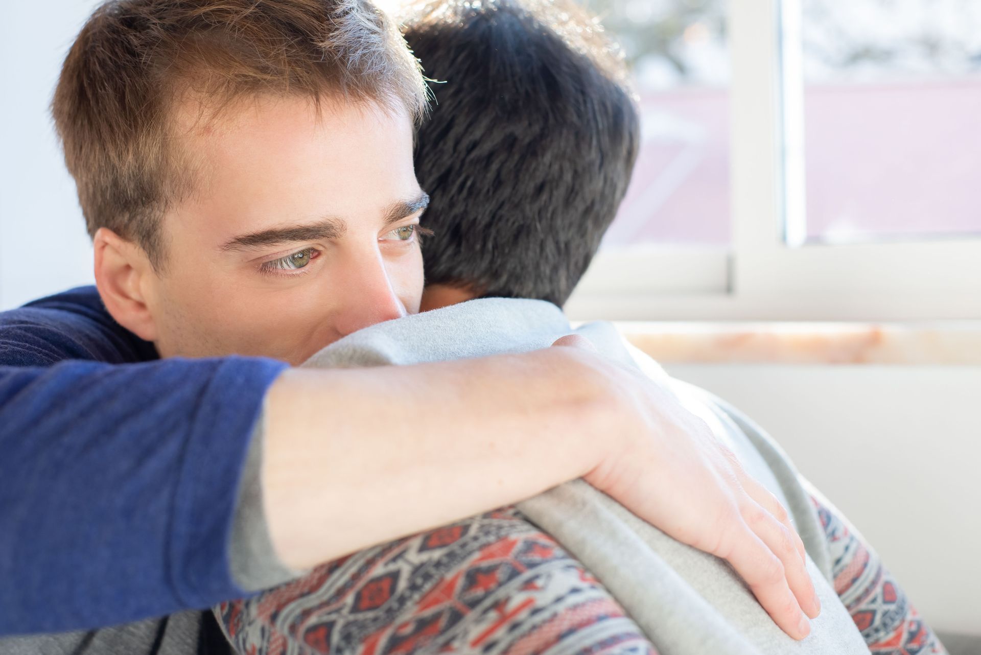 Two people embrace by a bright window, one resting their head on the other’s shoulder.