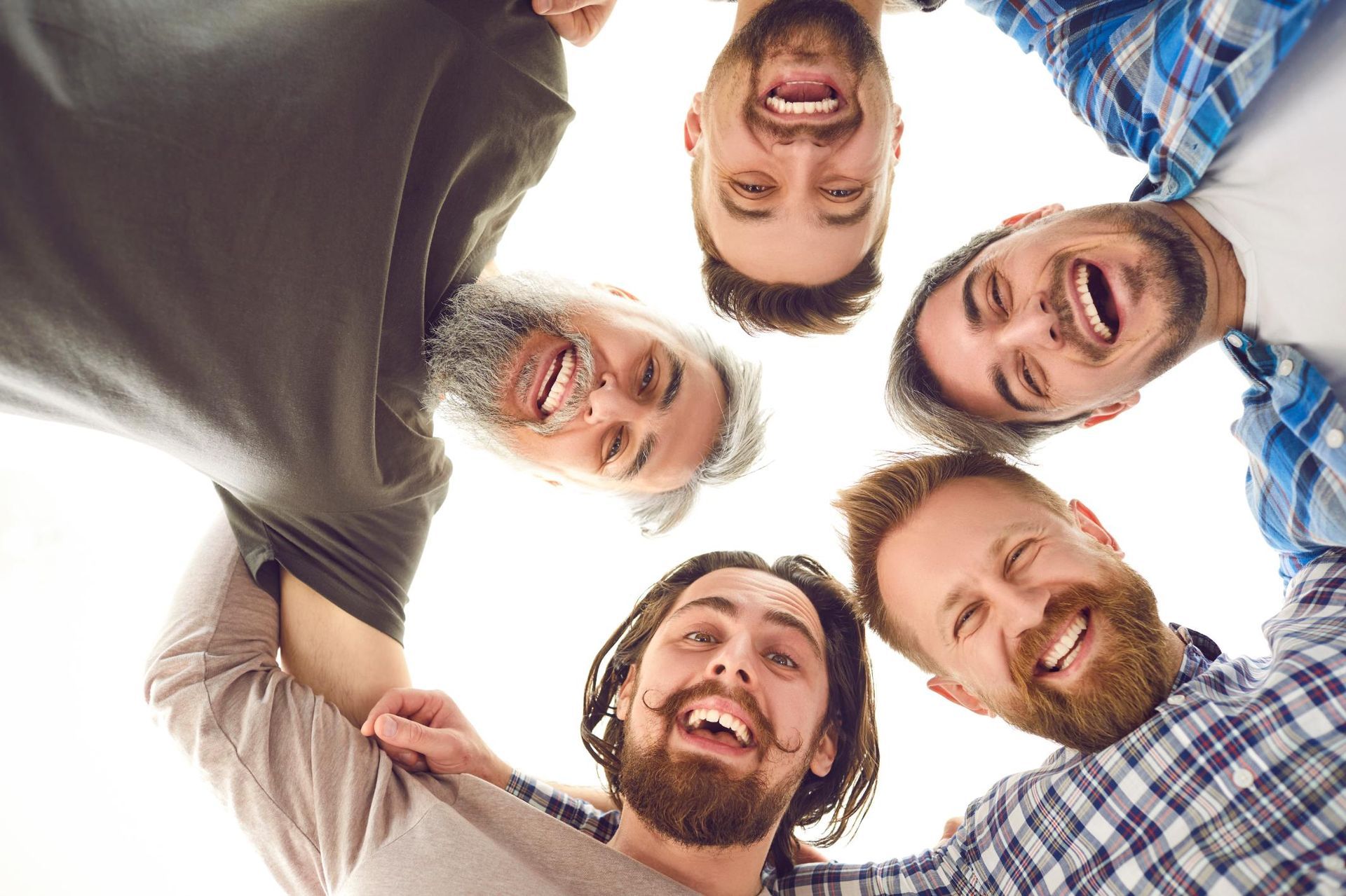 Five smiling friends leaning in a circle, looking down at the camera outdoors.
