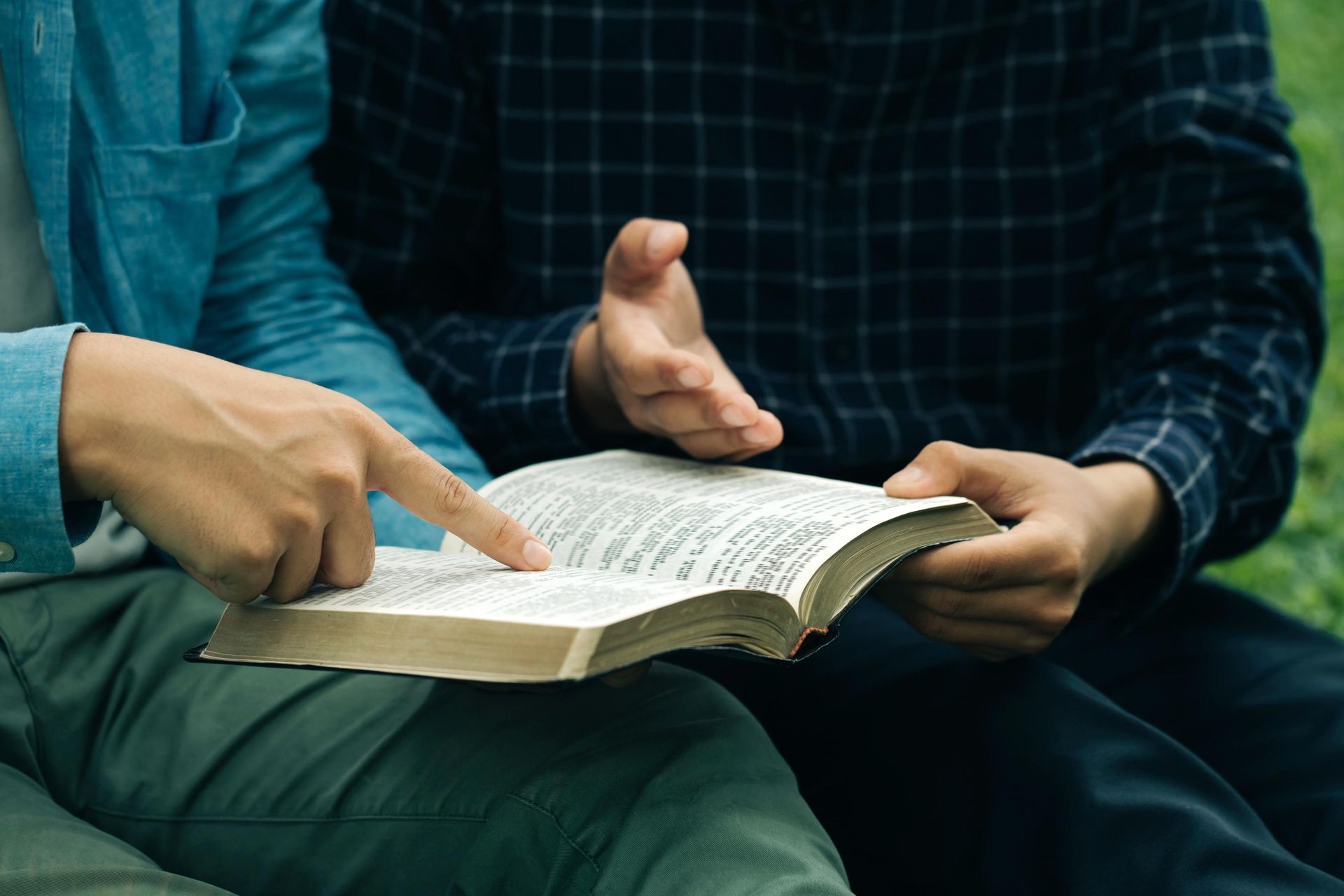 Two people seated outdoors, one pointing at an open book while the other holds it.