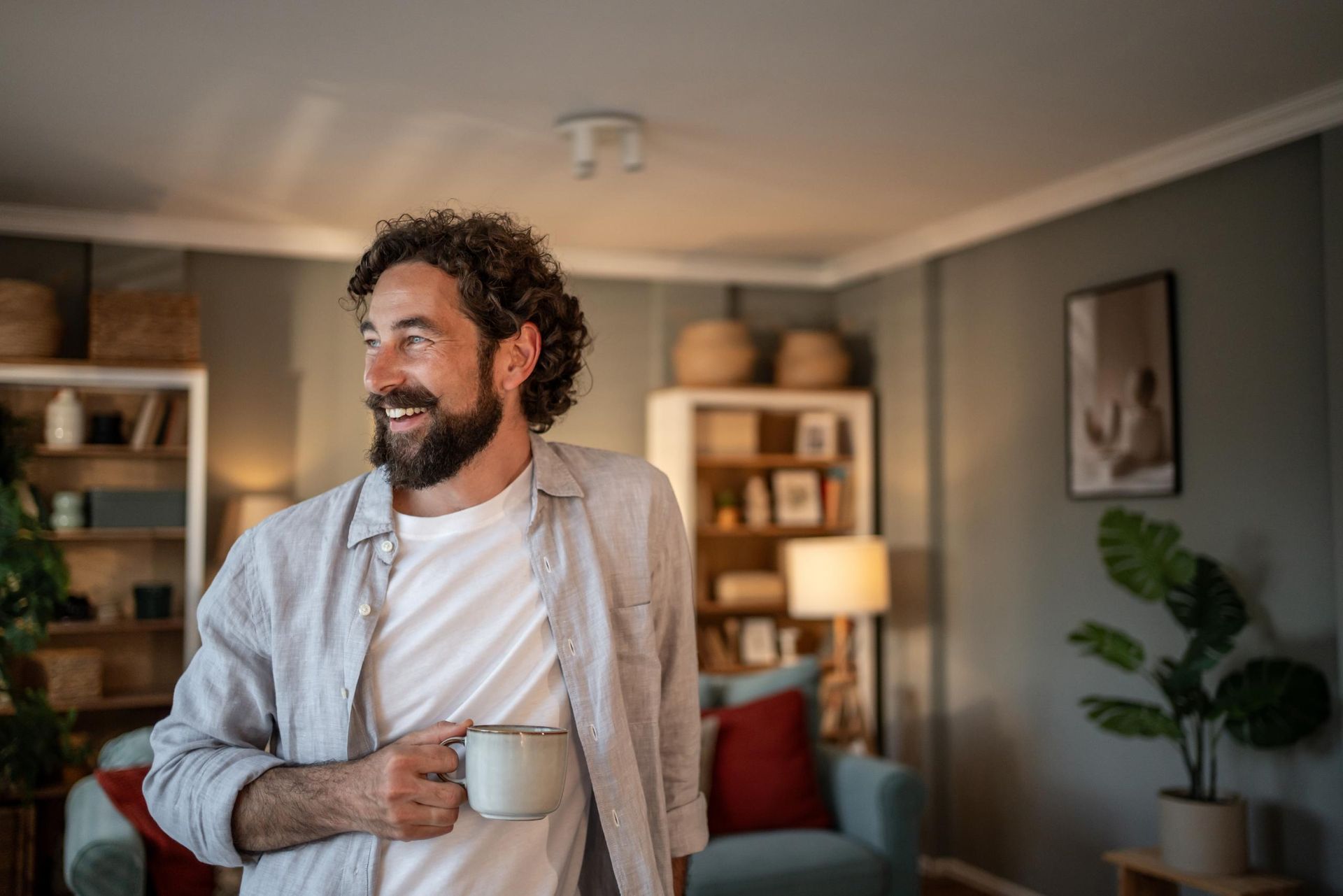 Smiling man holding a mug in a cozy living room with shelves, lamp, and plants