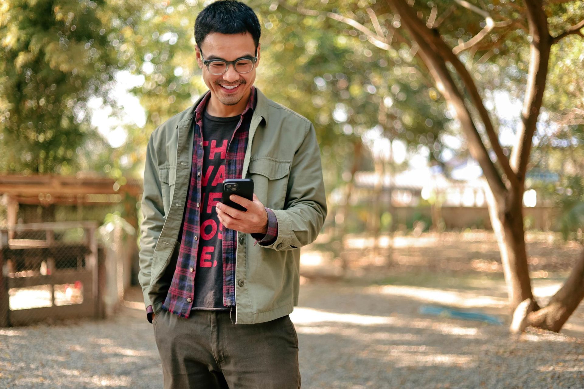 Person smiling at a phone in a sunlit park, wearing a green jacket and red plaid shirt.