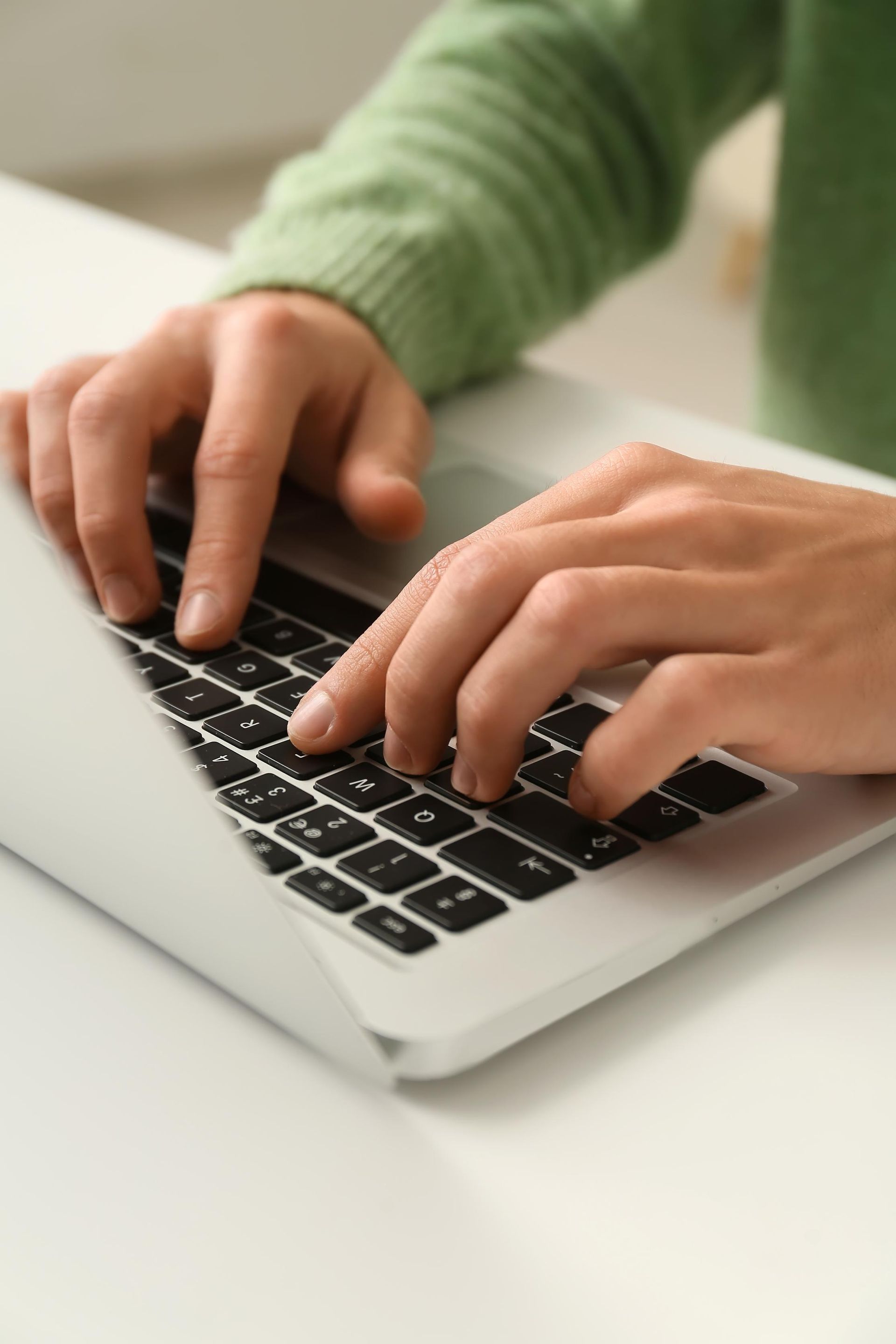 Hands typing on a laptop keyboard on a white desk, wearing a green sweater.