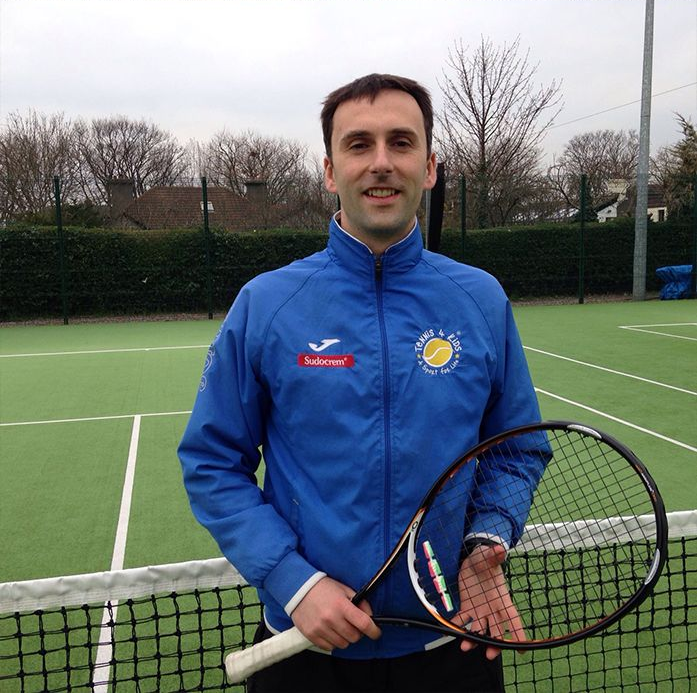Man holding a tennis racket on an outdoor court, wearing a blue jacket with a soccer club logo.