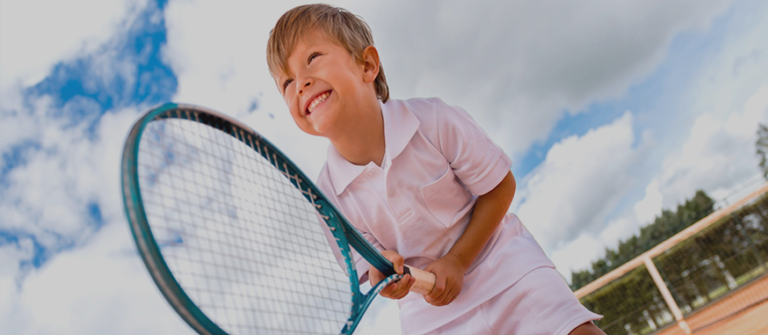 Smiling child holding a tennis racket on an outdoor court under a blue sky