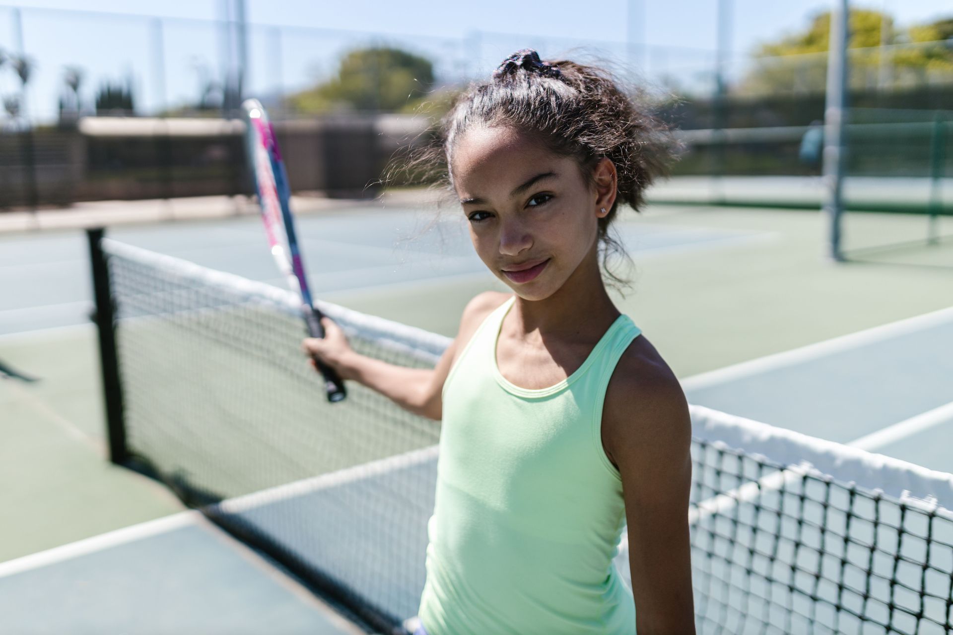 Person holding a tennis racket on an outdoor court, leaning on the net and smiling.