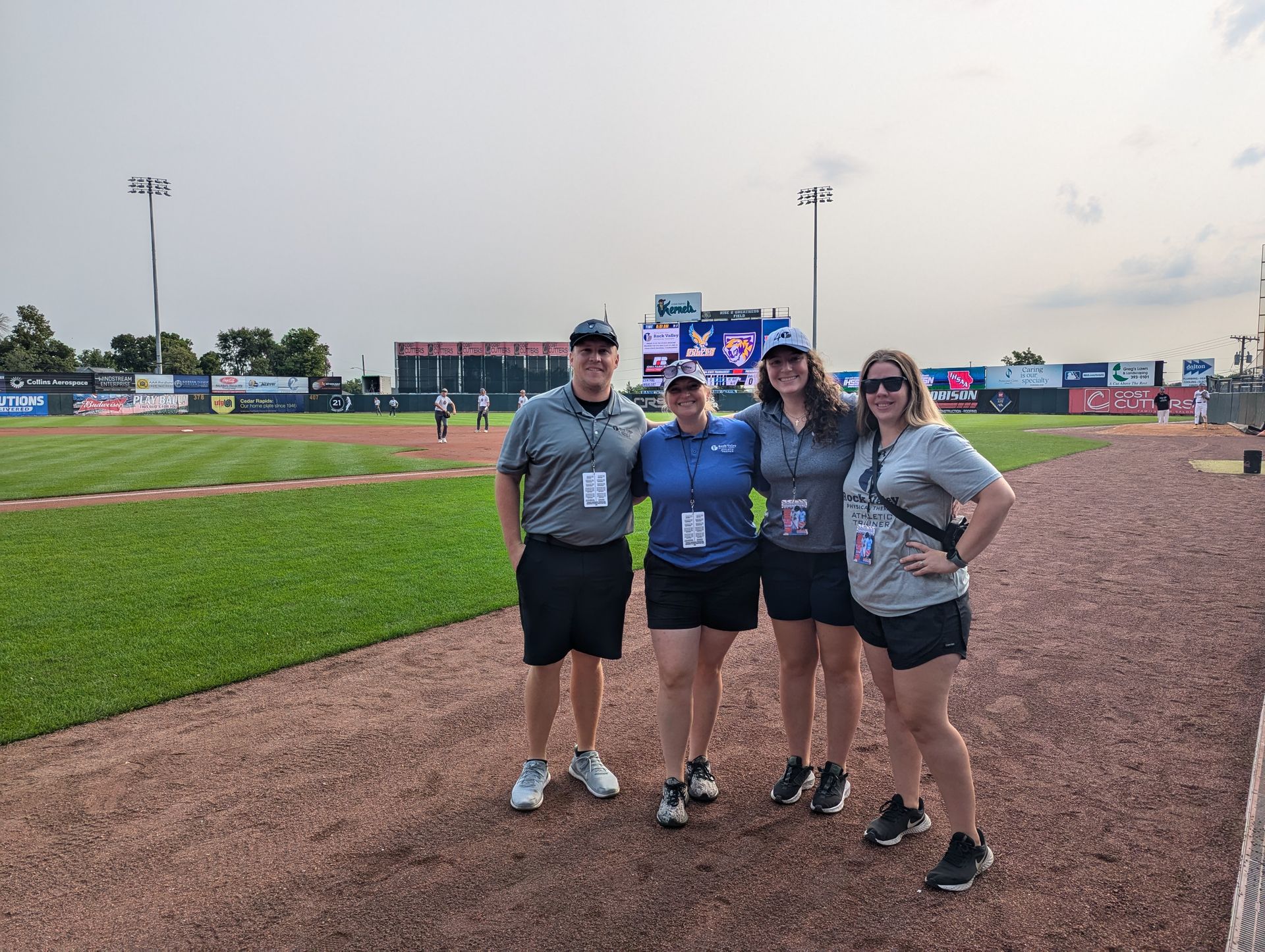 Four people posing on a baseball field, with grass, dirt infield, and stadium signs behind them.