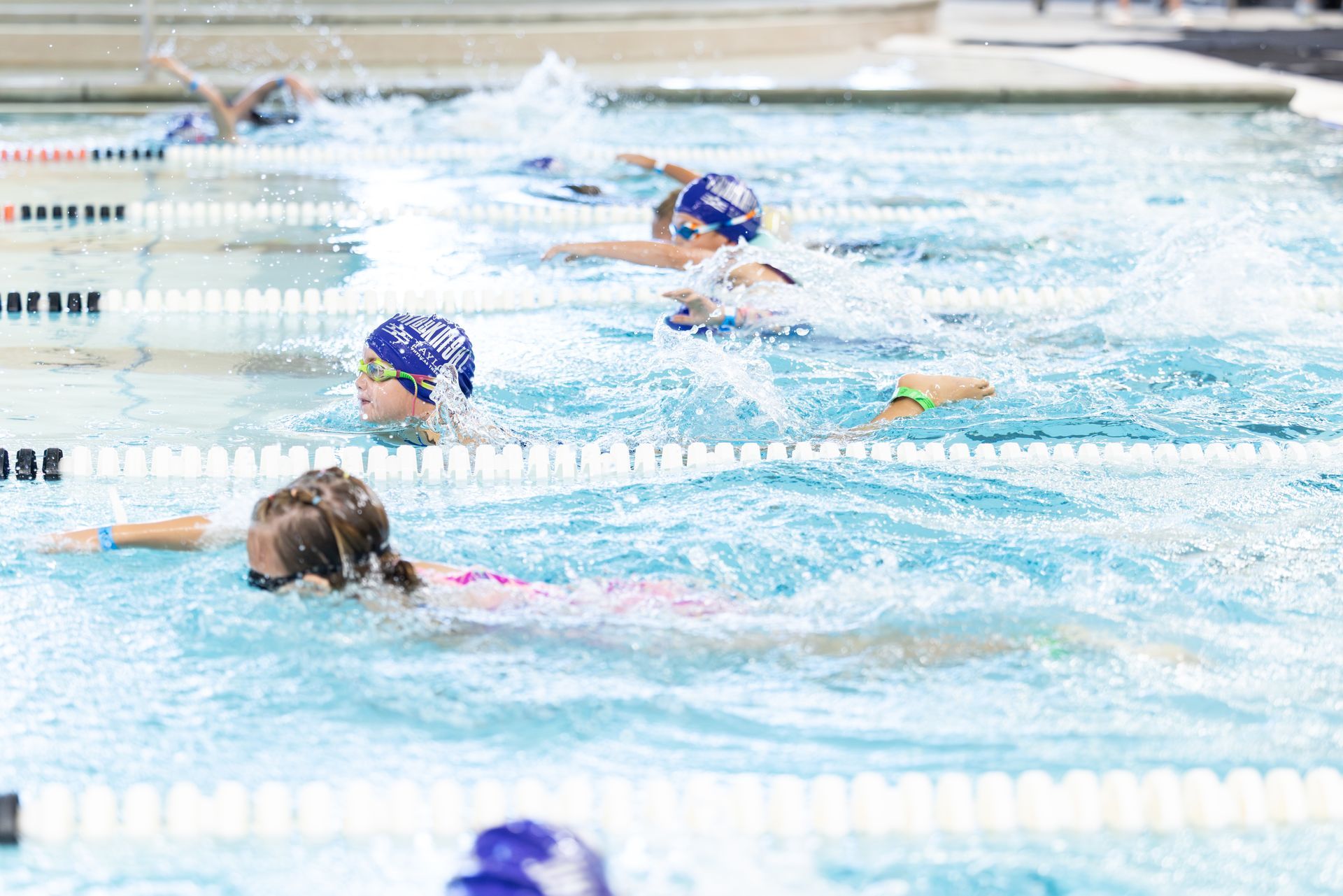 Swimmers in blue caps racing in an outdoor pool, water splashing as they swim laps.