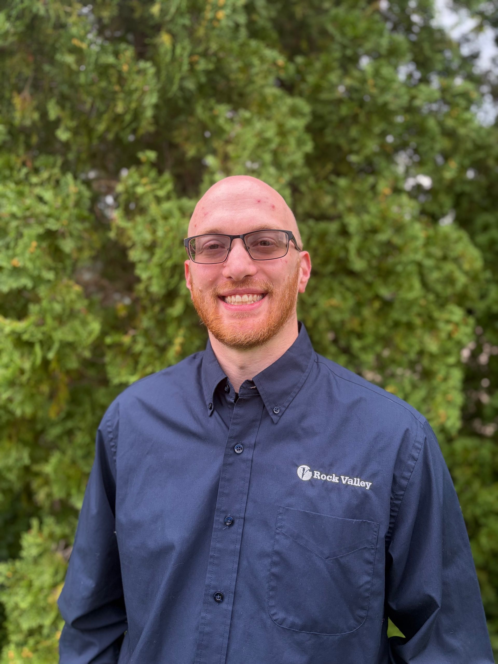 Man in navy shirt smiling outdoors with greenery in the background