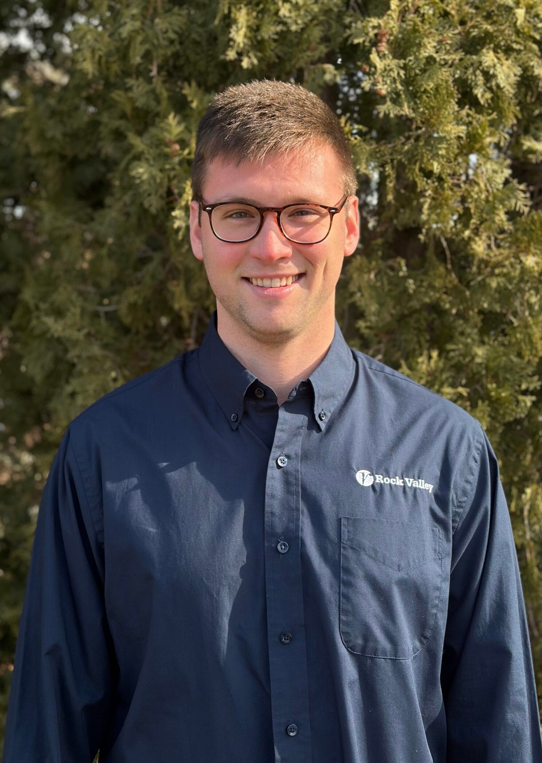 A person wearing glasses and a navy collared shirt with a logo, smiling in front of a tree.