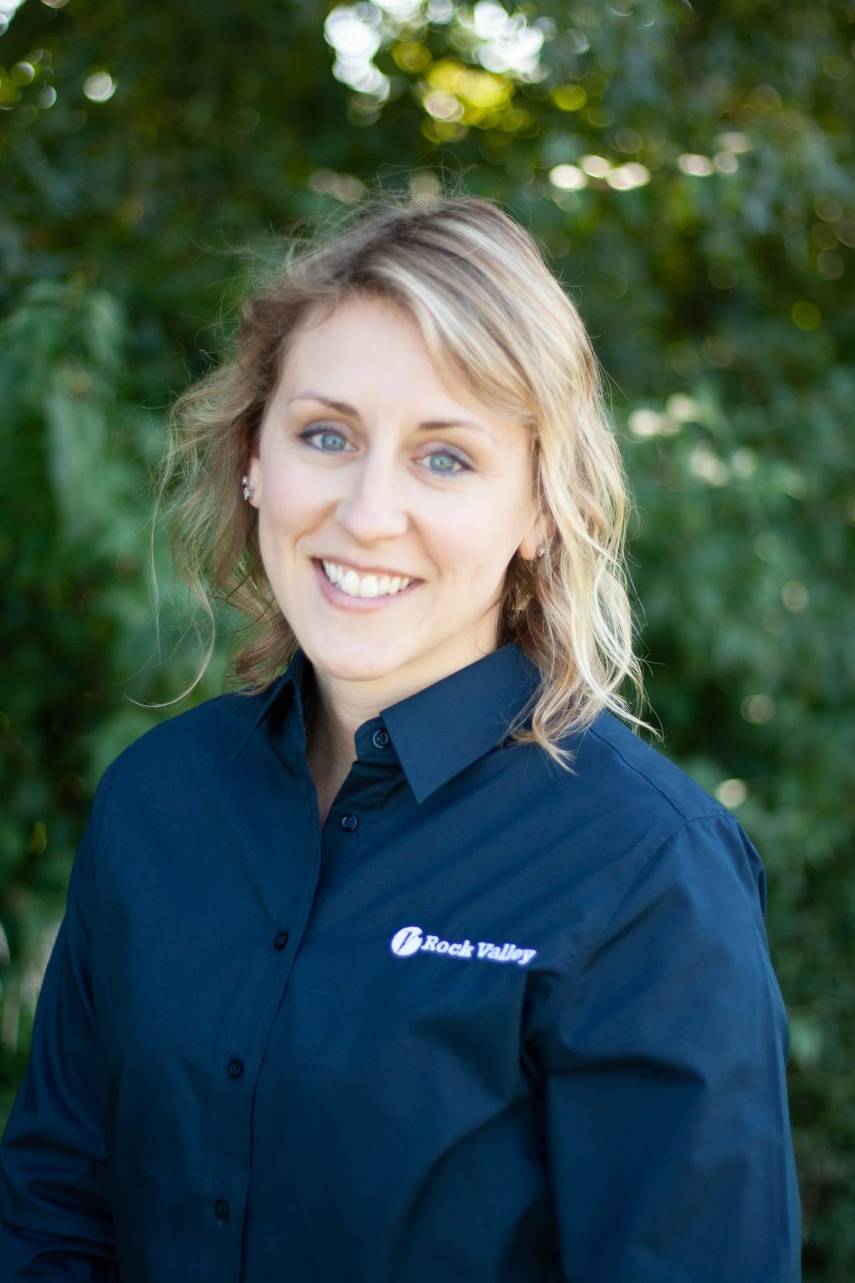 Smiling woman in a navy button-up shirt standing outdoors in front of green foliage