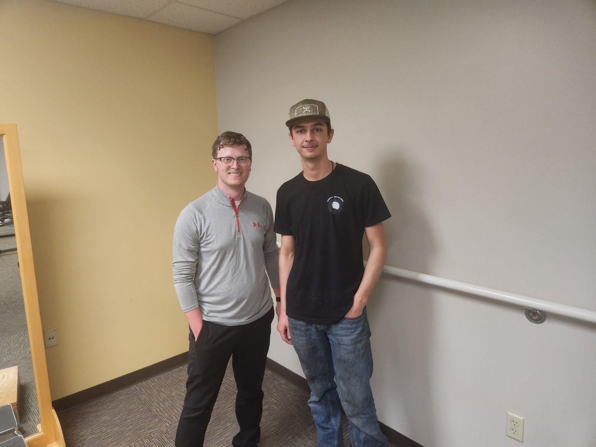 Two people standing in a hallway, one in a gray sweater and one in a black T-shirt and cap.